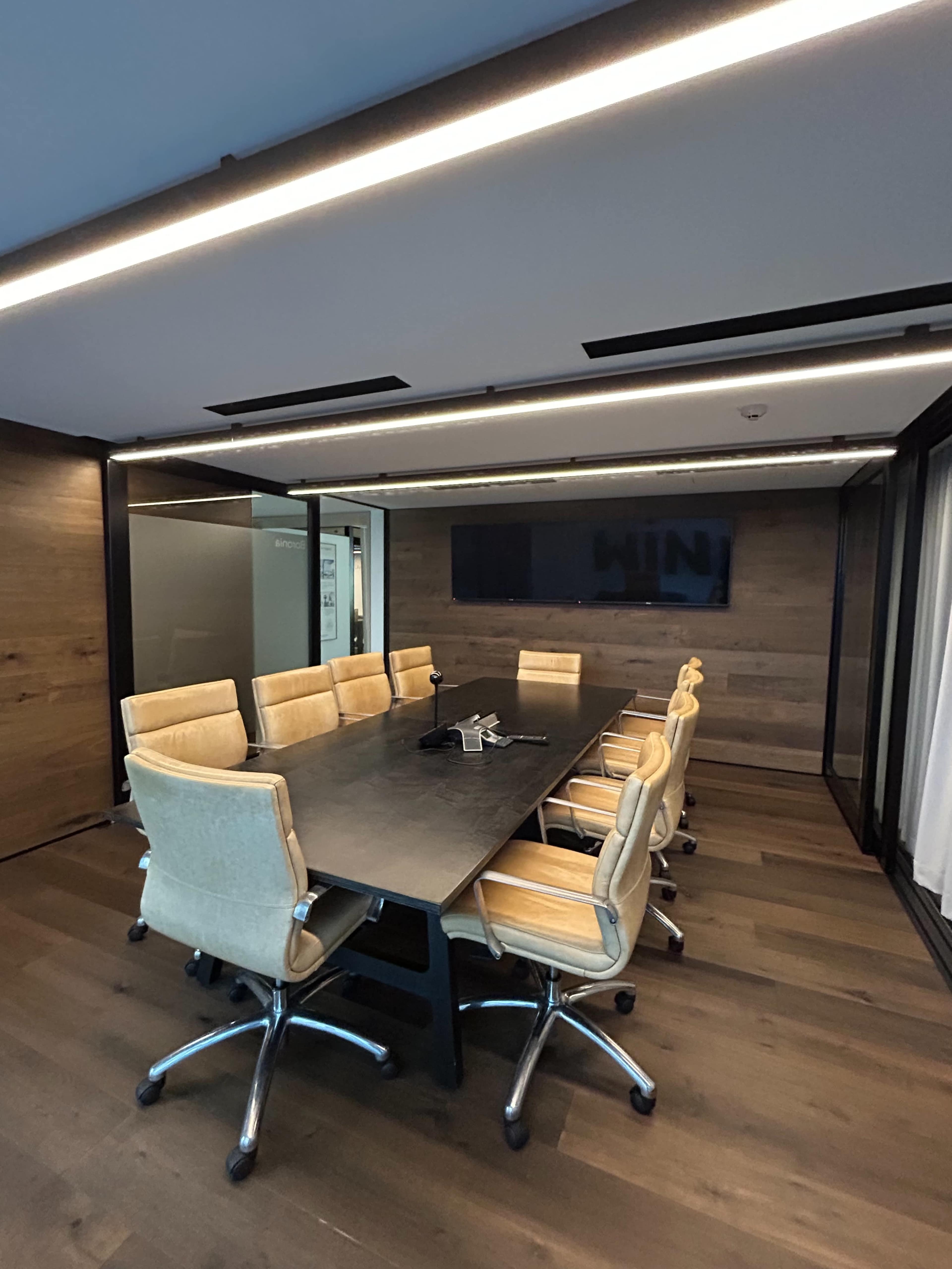 The image shows a modern conference room with a long black table surrounded by twelve beige chairs and a large wall-mounted screen.