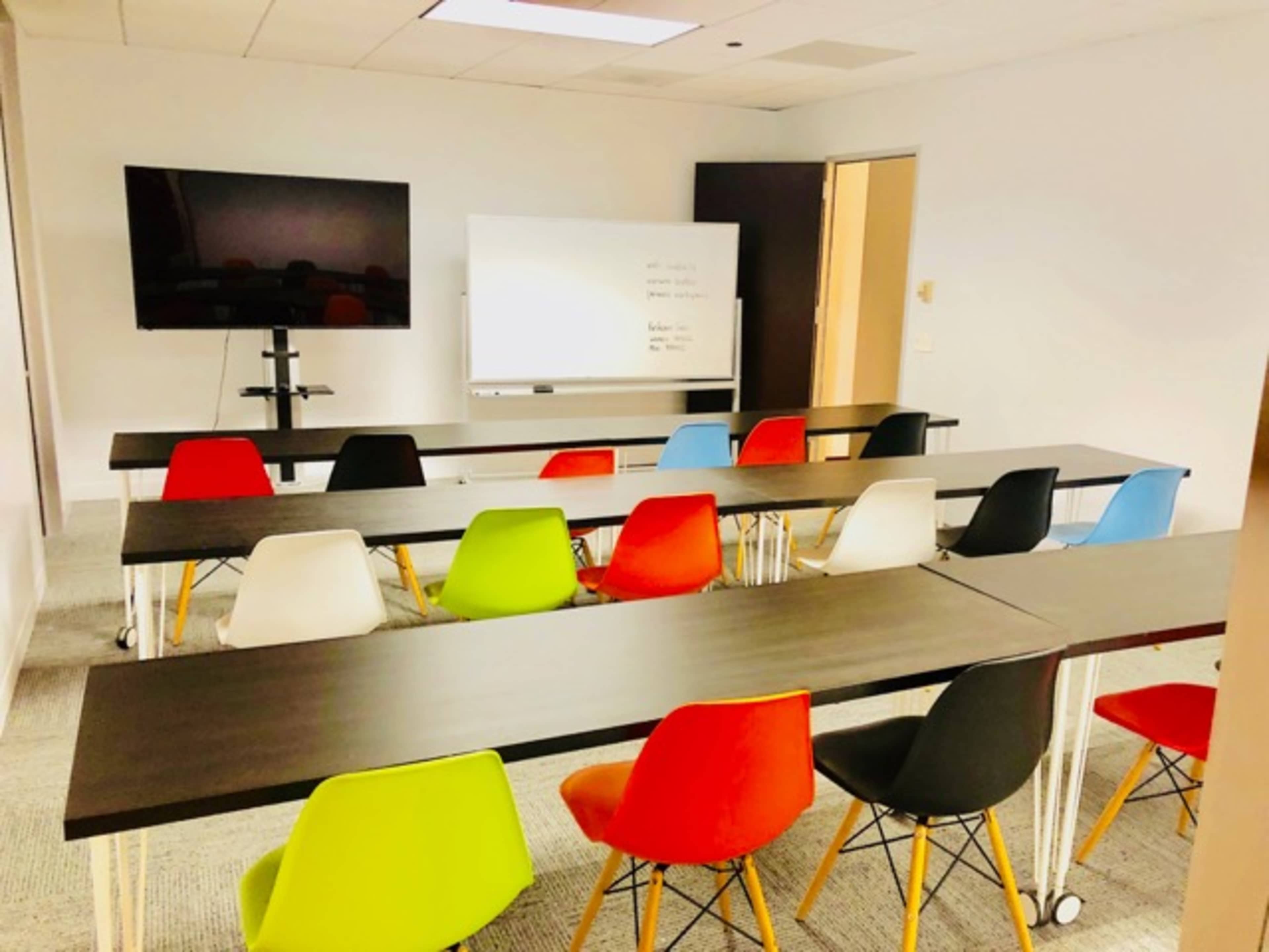 A conference room with several long tables arranged in rows, each accompanied by colorful chairs, and a wall-mounted television alongside a whiteboard.