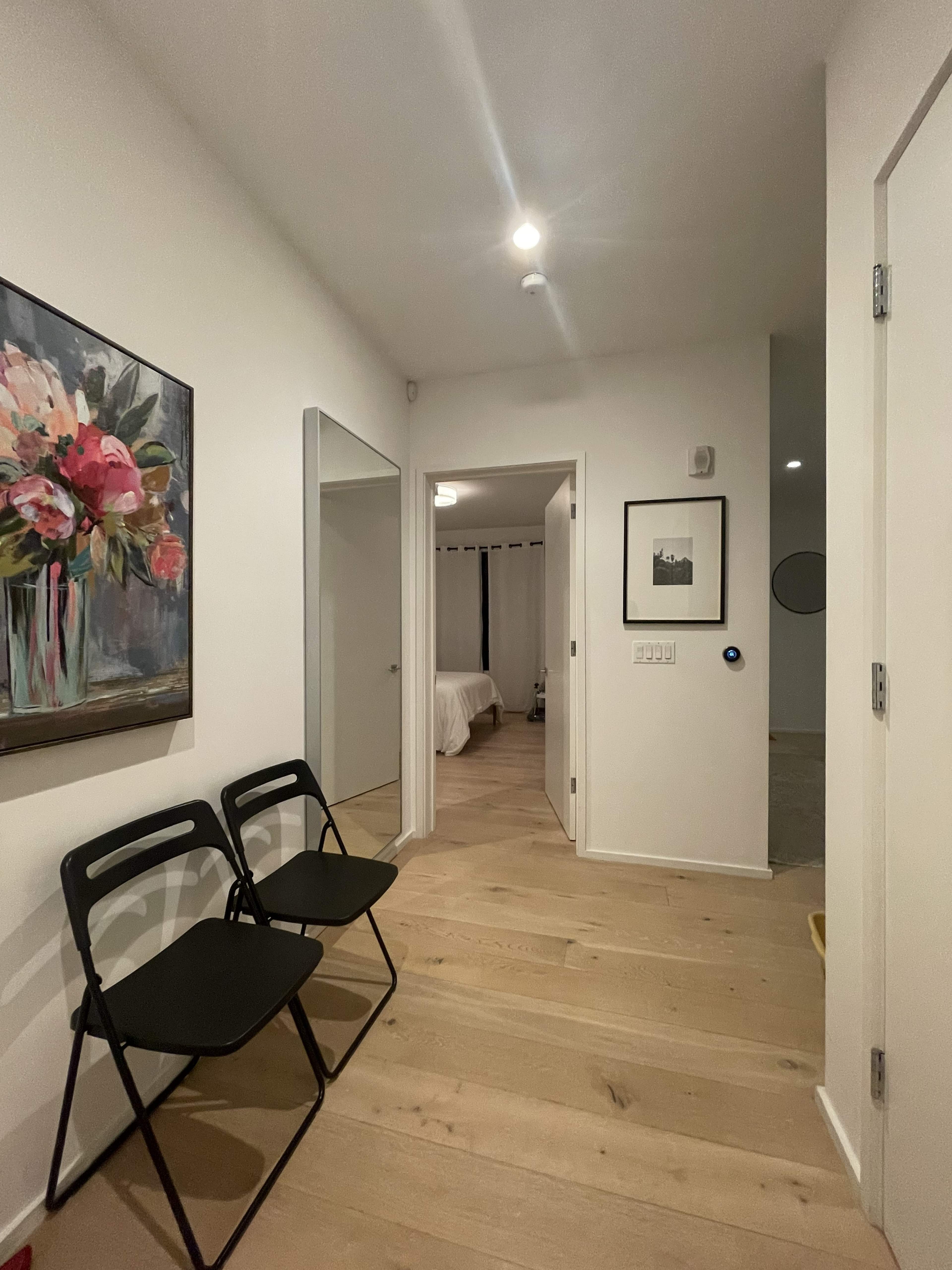 A narrow hallway with two black folding chairs, a floral painting on the wall, and a doorway leading to a bedroom.
