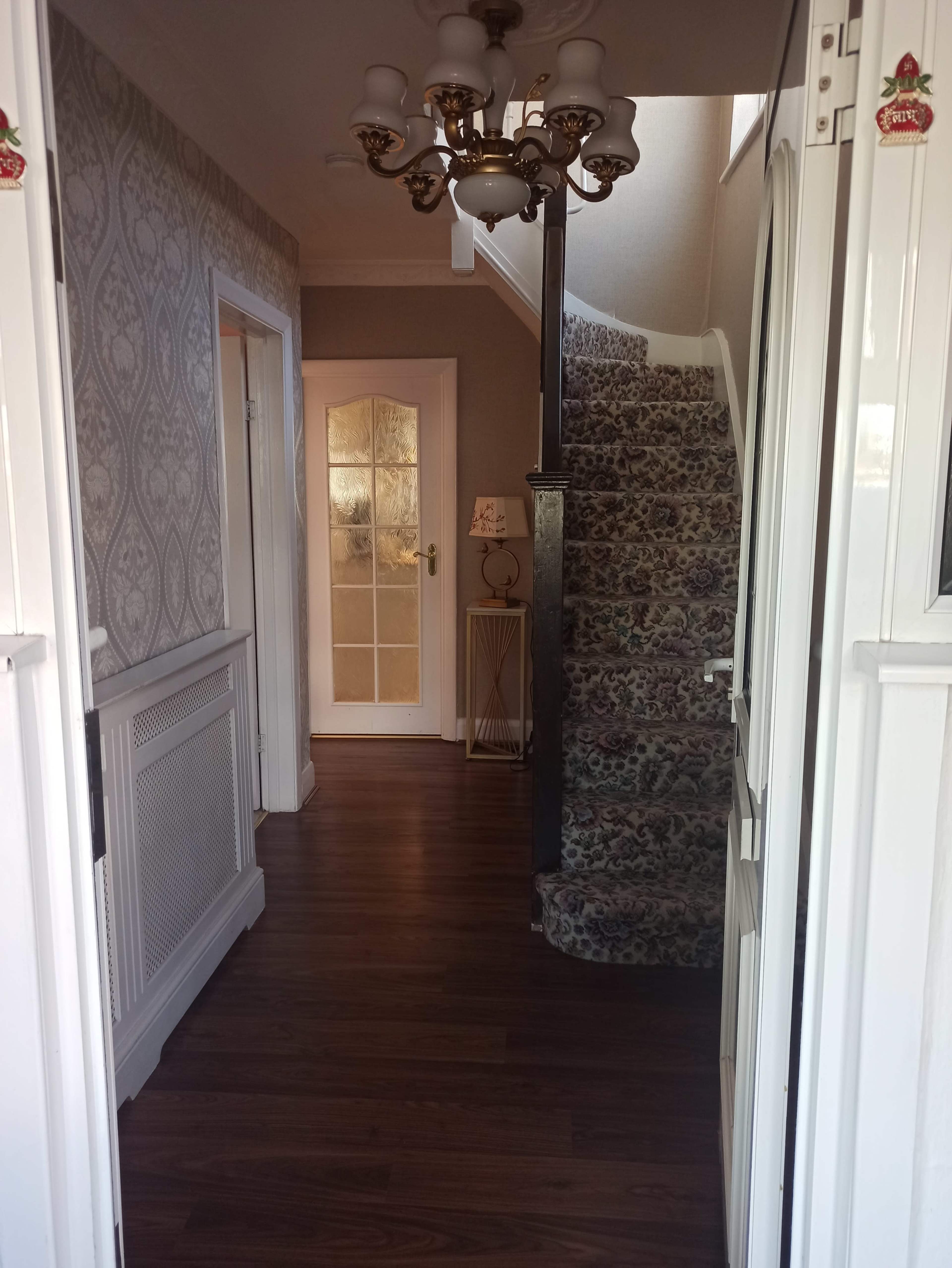 A hallway with a staircase leading upward, framed by wallpaper and wooden flooring, with a door at the end and light fixtures above.