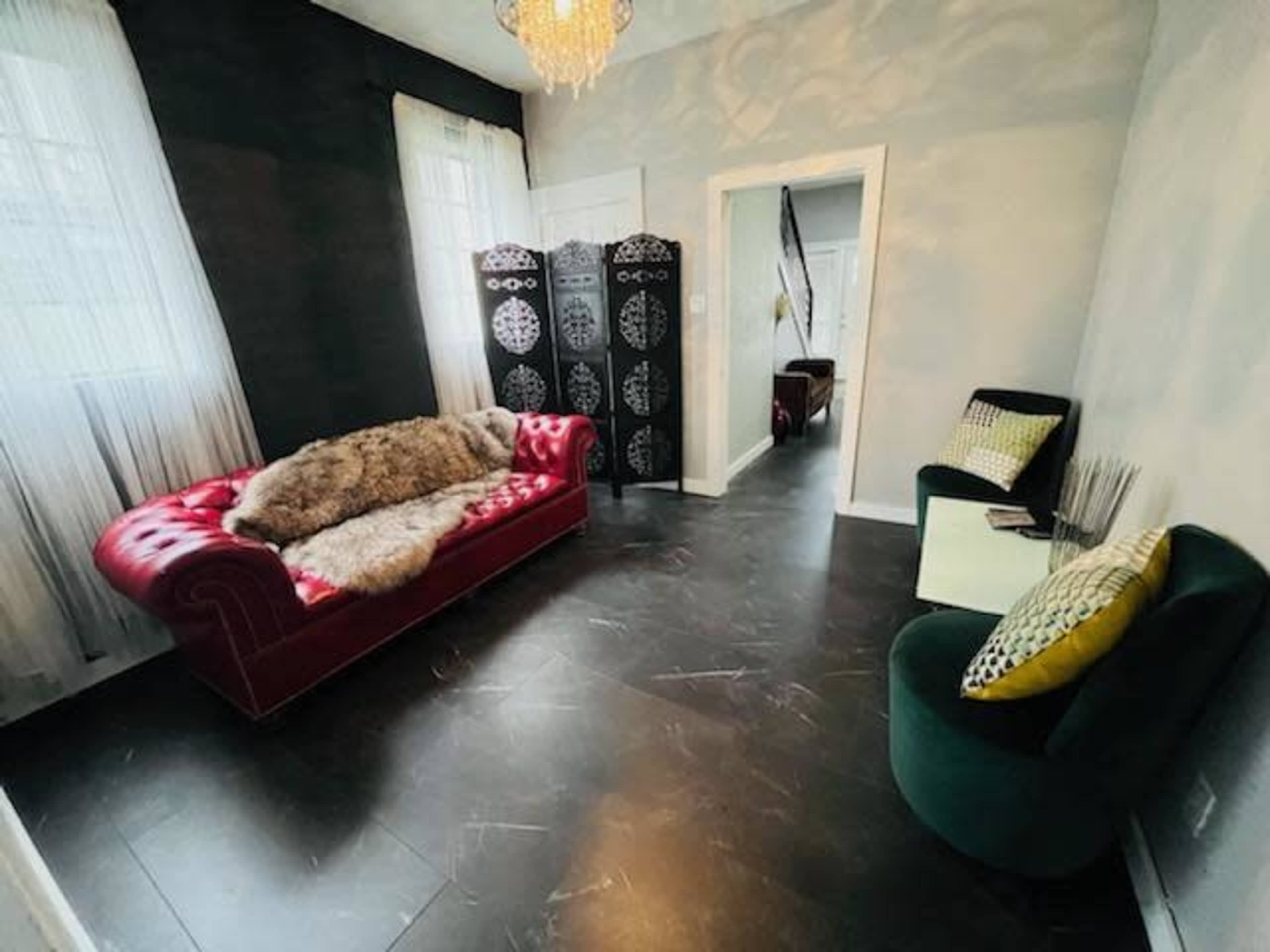 The image shows a room with a red velvet sofa, a fur throw, two chairs, and a decorative screen, illuminated by natural light through sheer curtains.