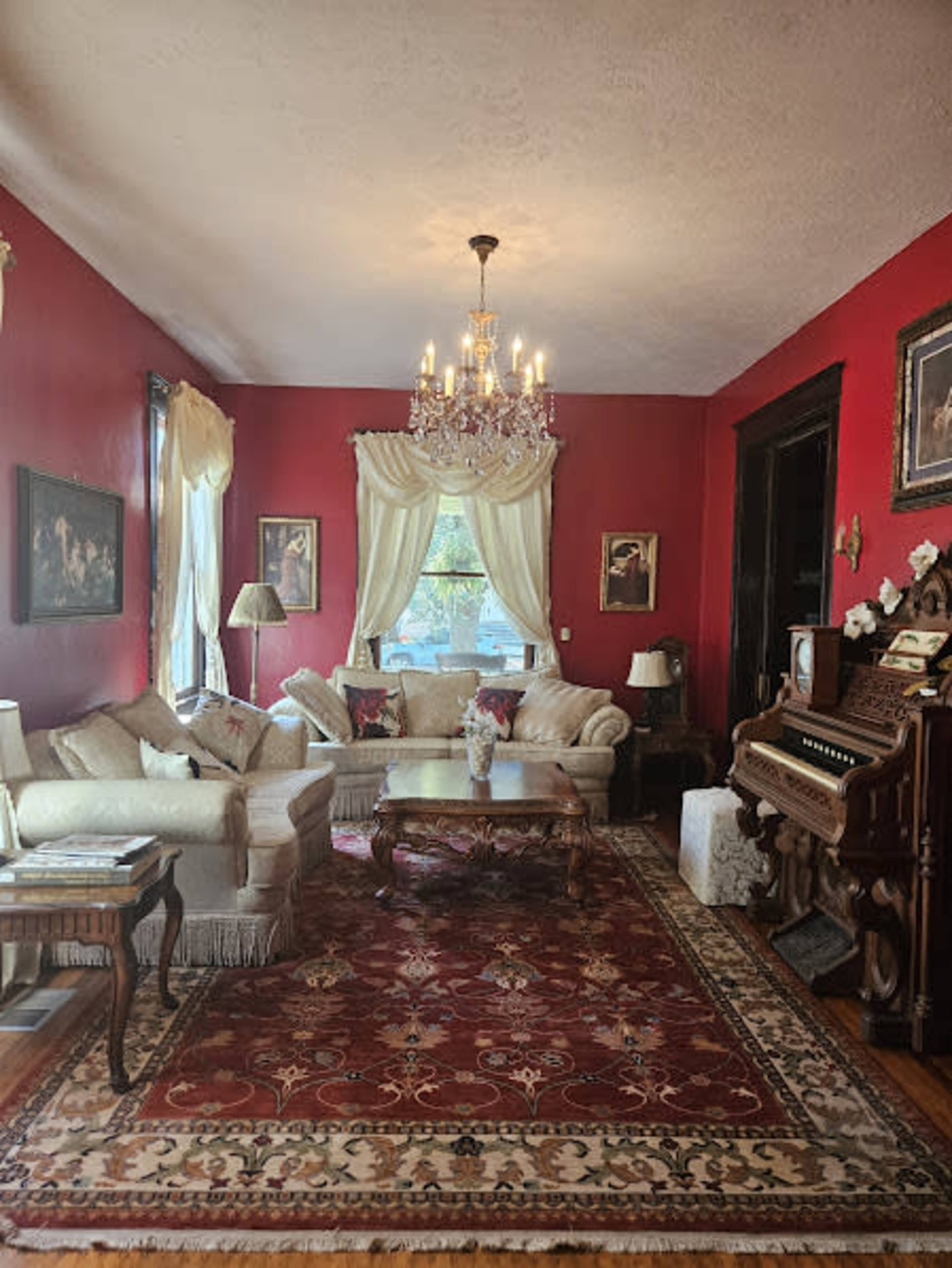 The living room features red walls, a chandelier, a large area rug, and a mix of vintage furniture, including a piano.