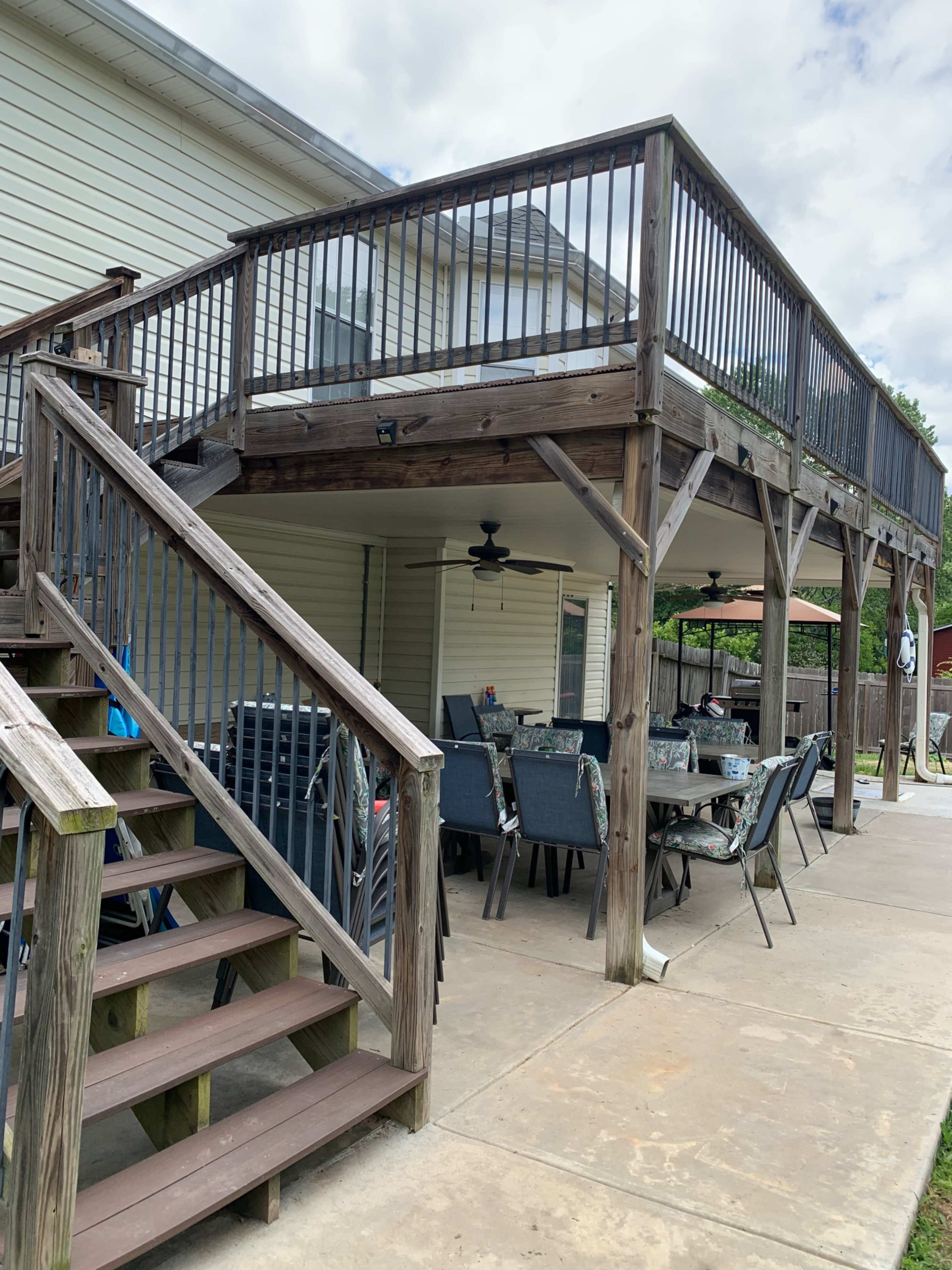 The image shows a wooden deck elevated above a concrete patio, with a table set for dining and a staircase leading down to the ground level.