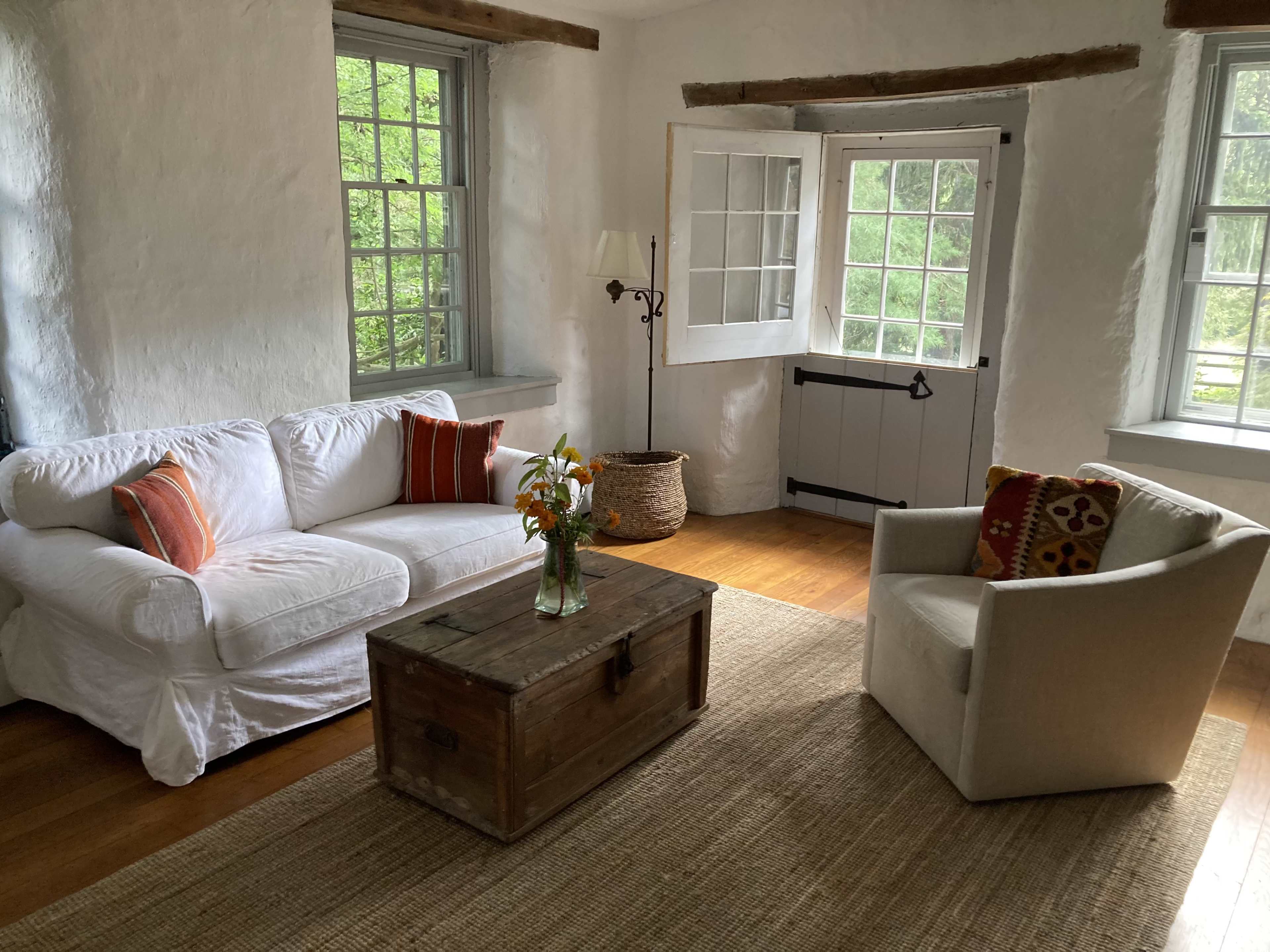 The room features a white sofa with decorative pillows, a wooden coffee table, and a beige armchair, all set against white walls and large windows that open to a green view.