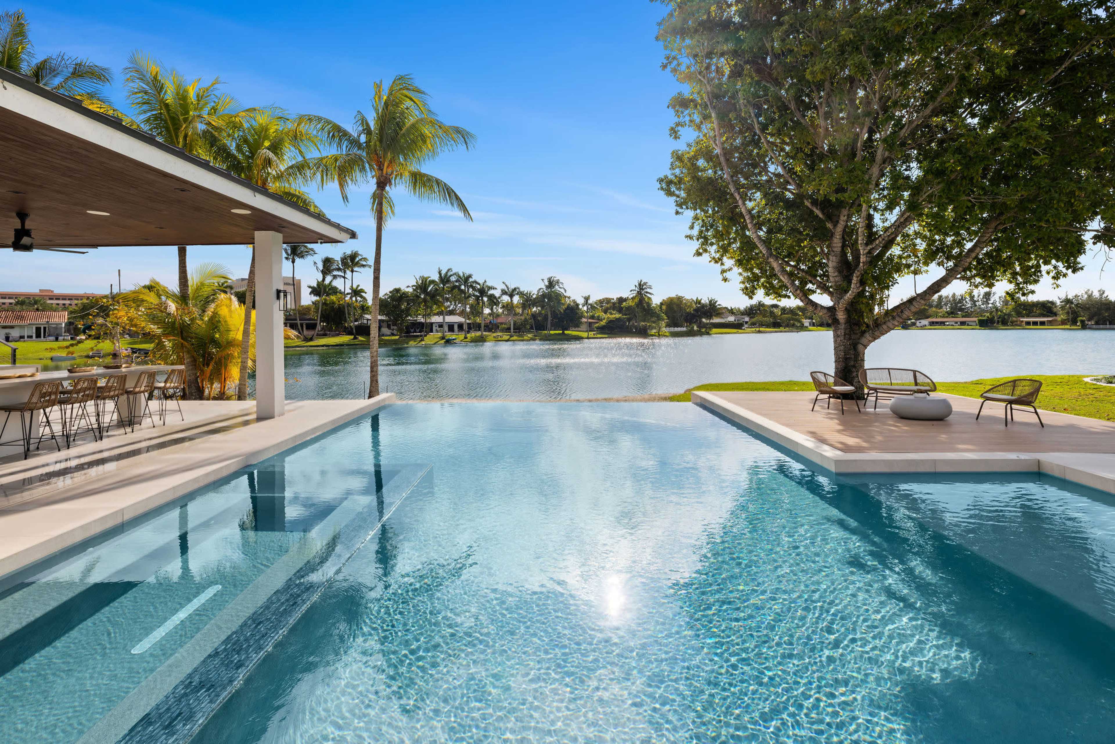 The image shows a modern swimming pool adjacent to a waterfront, surrounded by palm trees and a spacious outdoor dining area.