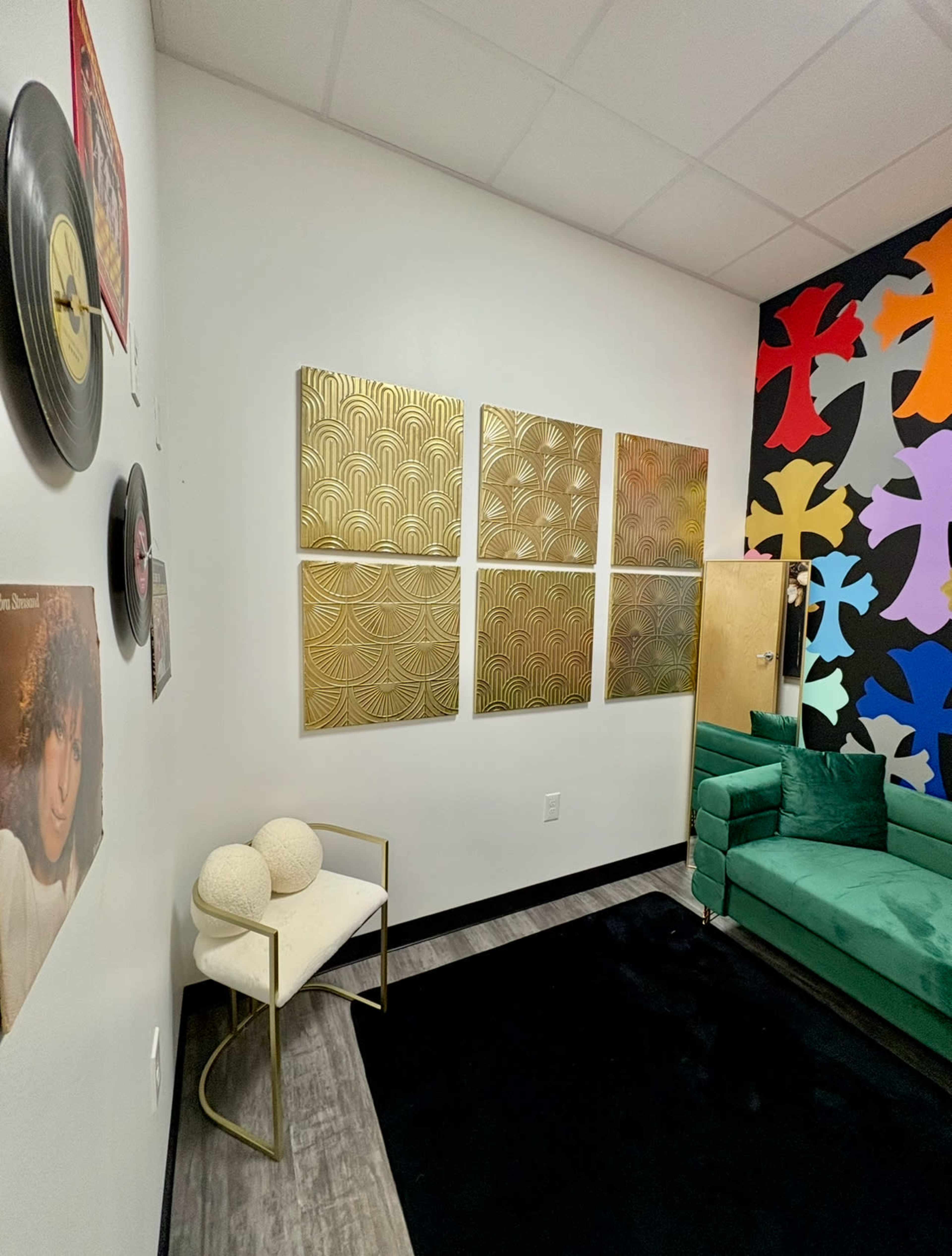 The image shows a modern lounge area with a green couch, a black rug, and decorative gold wall art, alongside a vinyl record and a colorful mural on the wall.