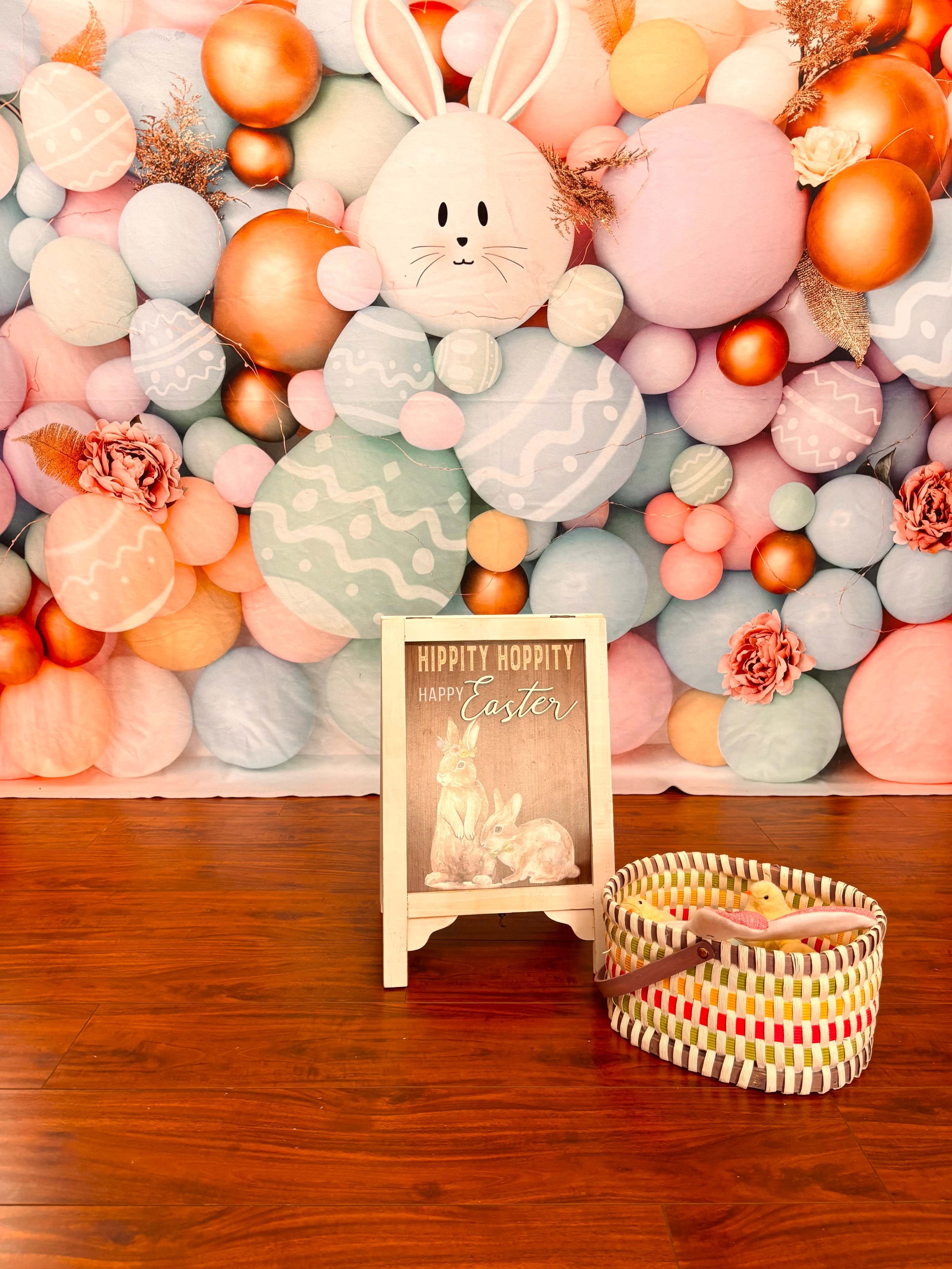 The image features a decorative backdrop of colorful balloons and a sign that reads "HIPPITY HOPPITY HAPPY EASTER," accompanied by a basket.
