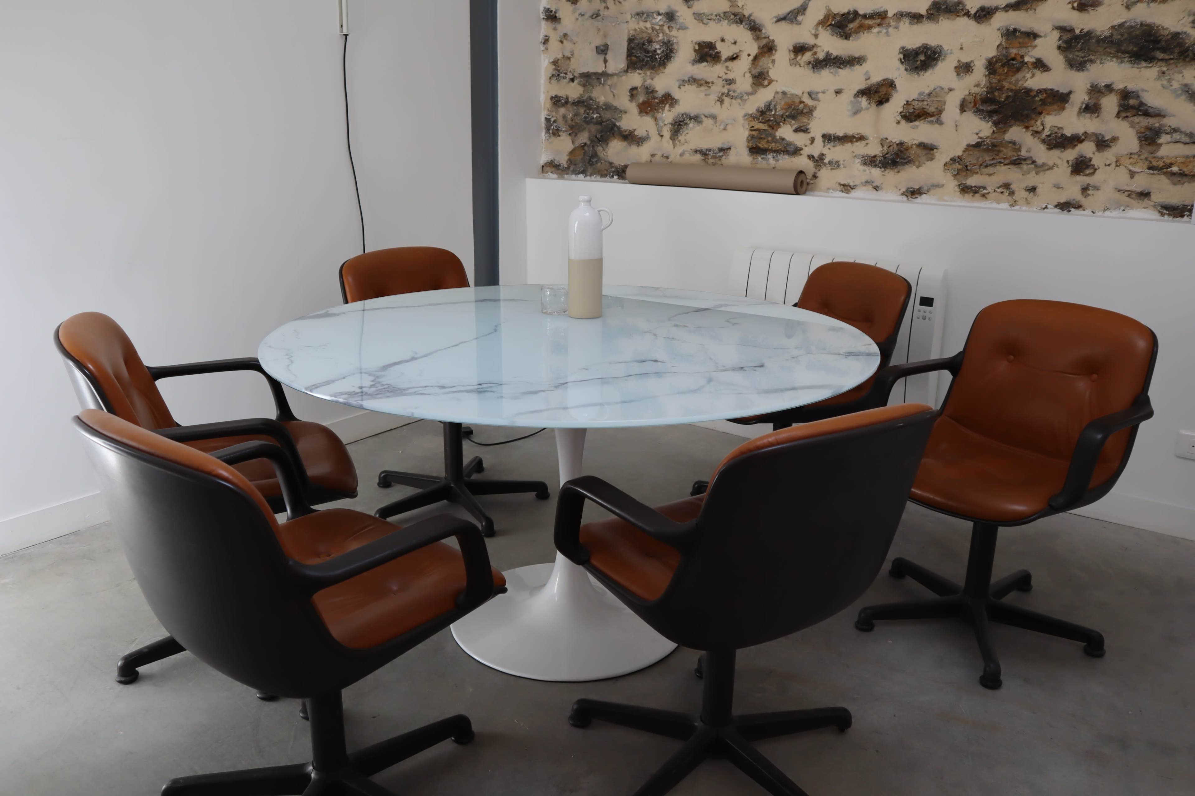 A round marble table is surrounded by five rolling office chairs in a room with a stone wall.