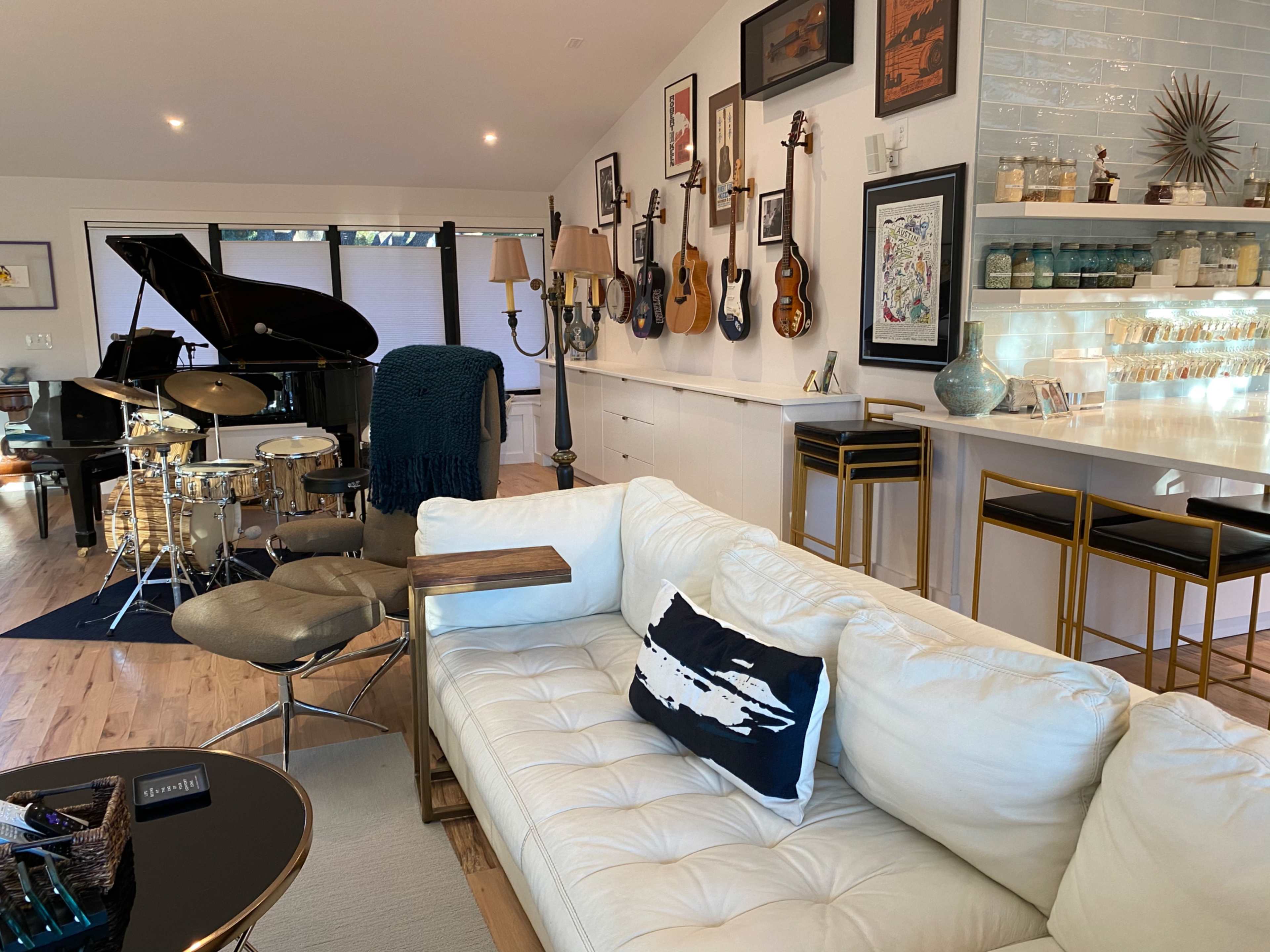 The image shows a modern living space featuring a white leather sofa, a grand piano, a drum set, various guitars on the wall, and a bar area with decorative items.