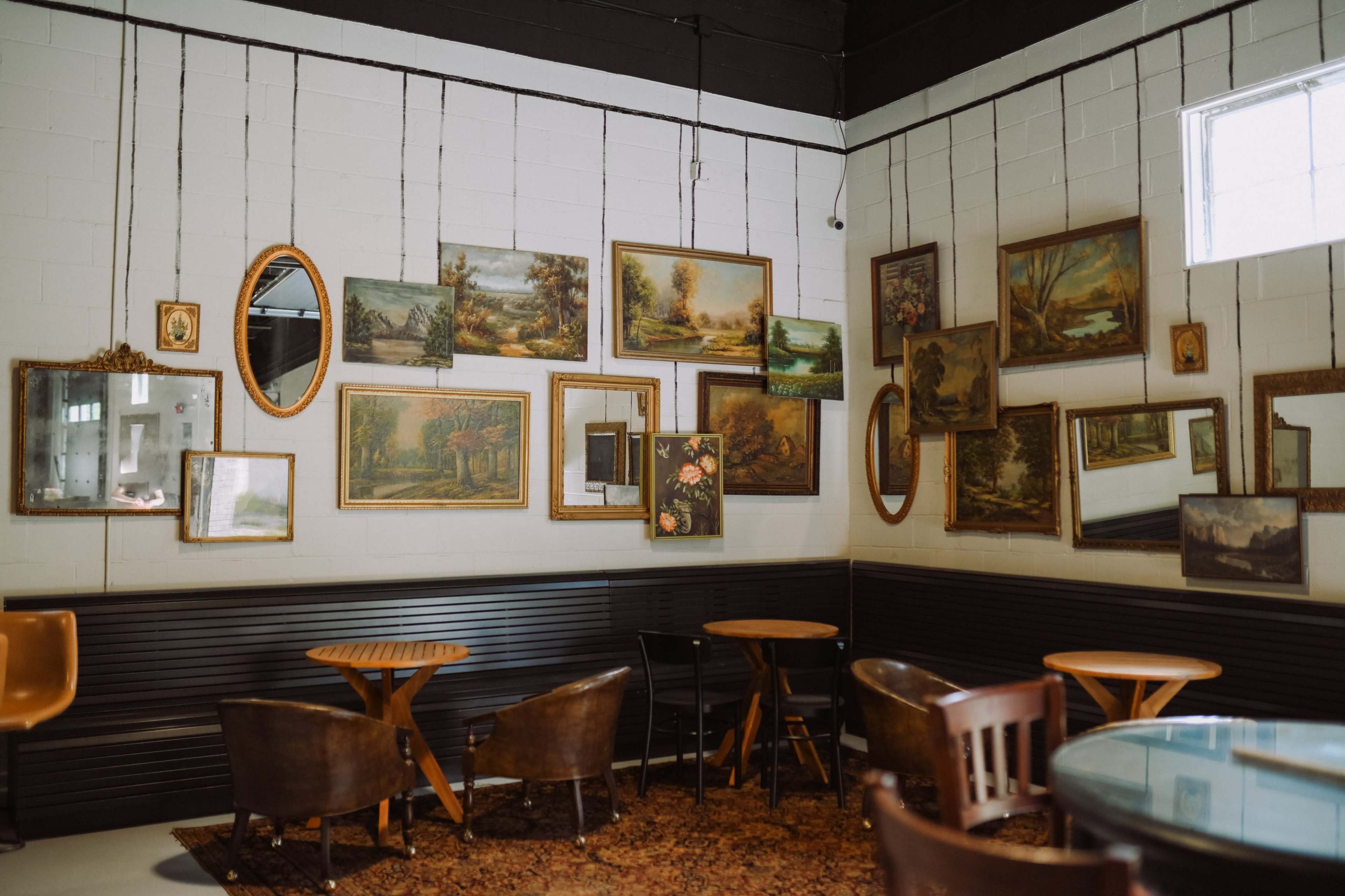 The image shows a cafe interior featuring several framed paintings and mirrors hanging on a white wall, with wooden tables and chairs arranged nearby.
