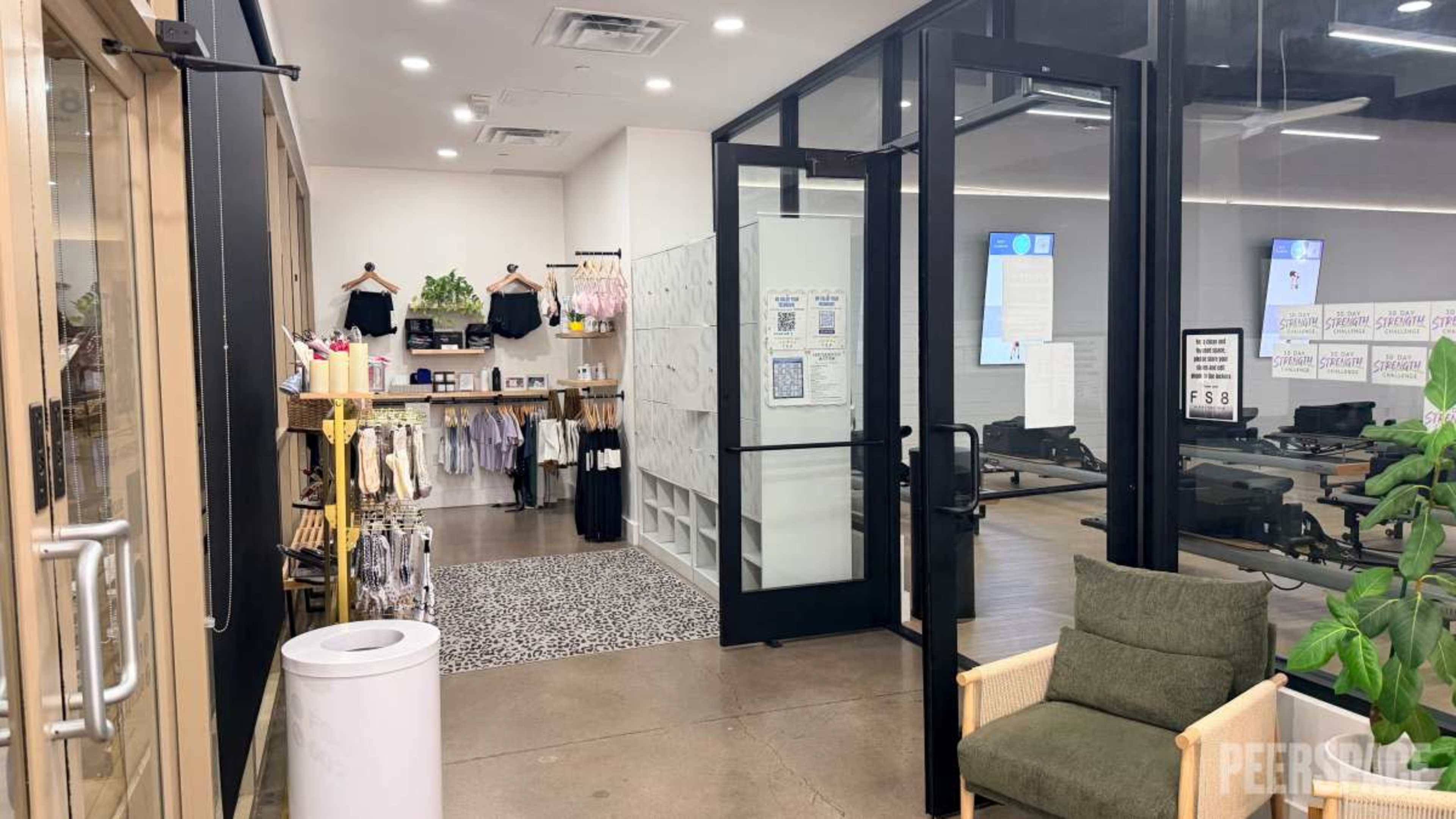 The image shows the interior of a fitness studio with a reception area featuring clothing racks, a seating area, and workout equipment visible in the background.