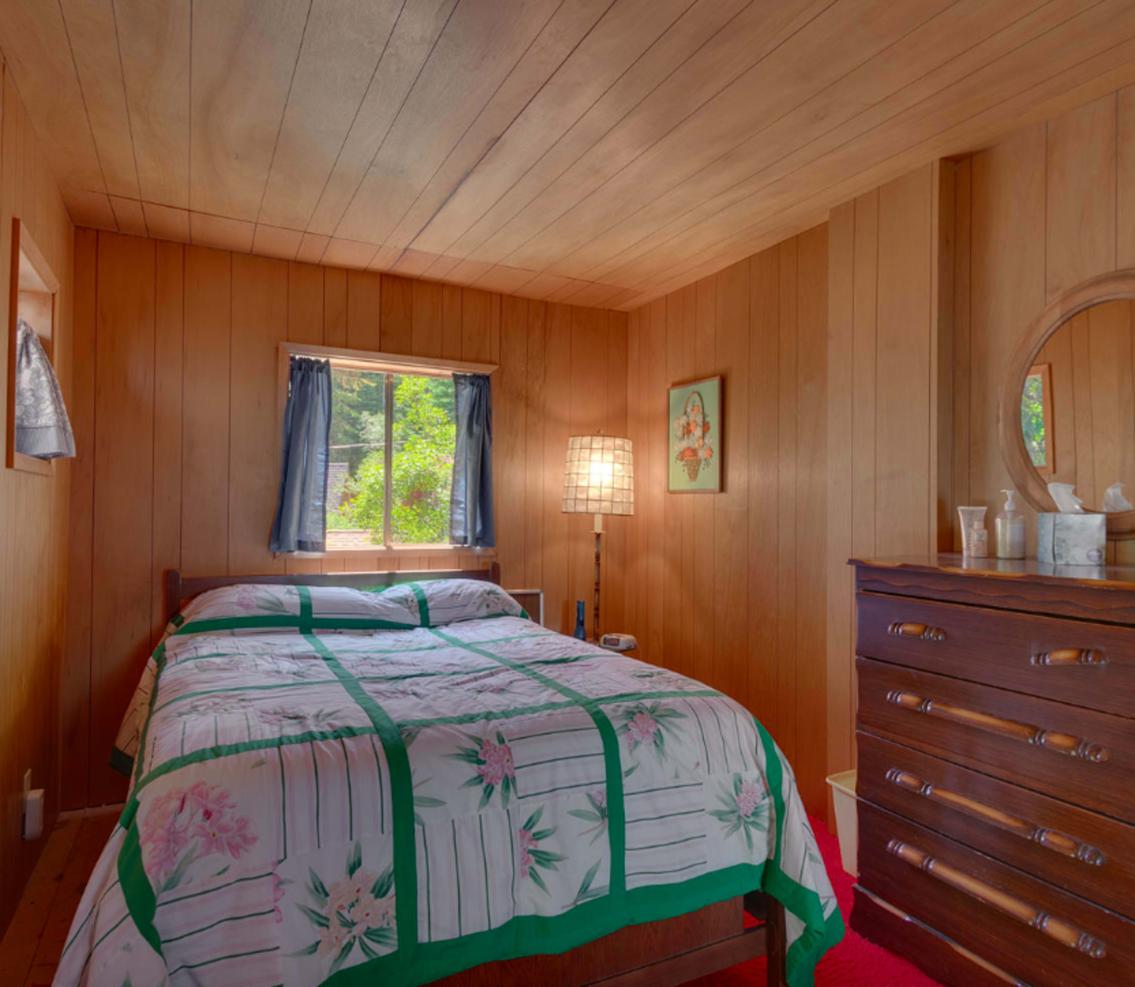 A small wooden bedroom featuring a bed with a floral-patterned quilt, a bedside table, a wooden dresser, and a window with curtains.