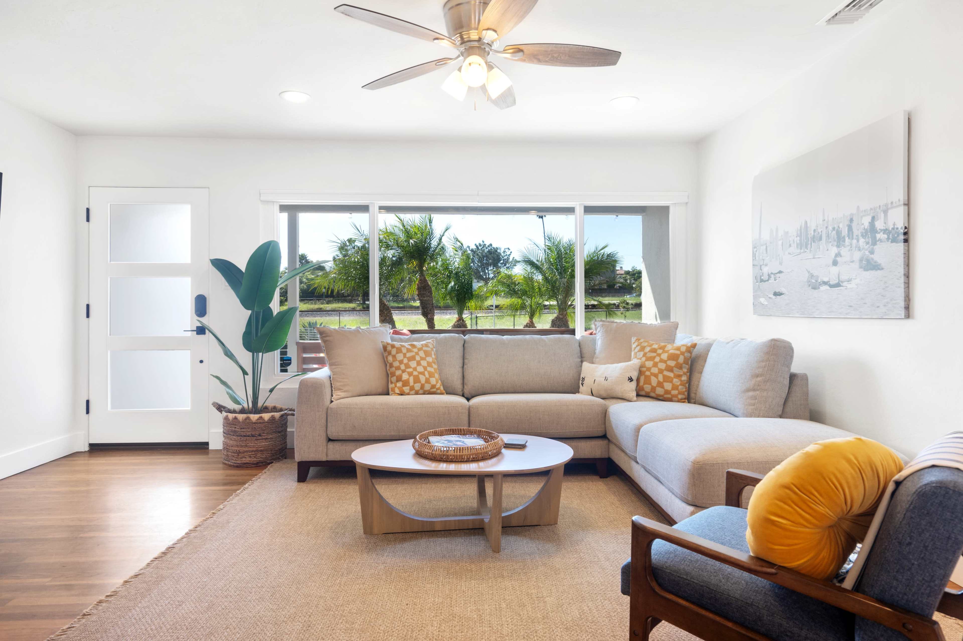 A bright living room features a sectional sofa, a round coffee table, and large windows overlooking a landscaped outdoor area with palm trees.