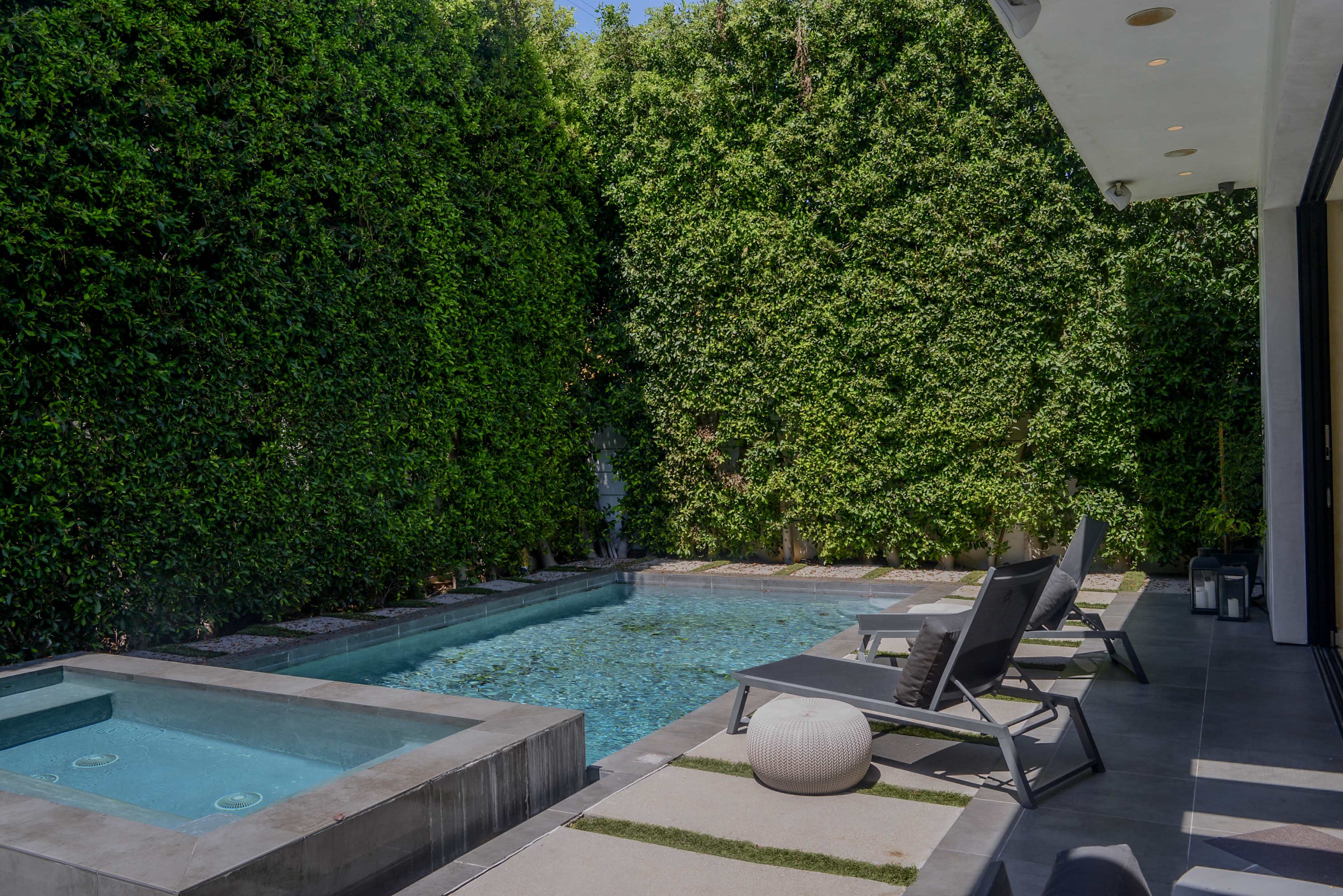 The image features a modern backyard with a swimming pool, spa, and lounge chairs surrounded by lush green hedges.