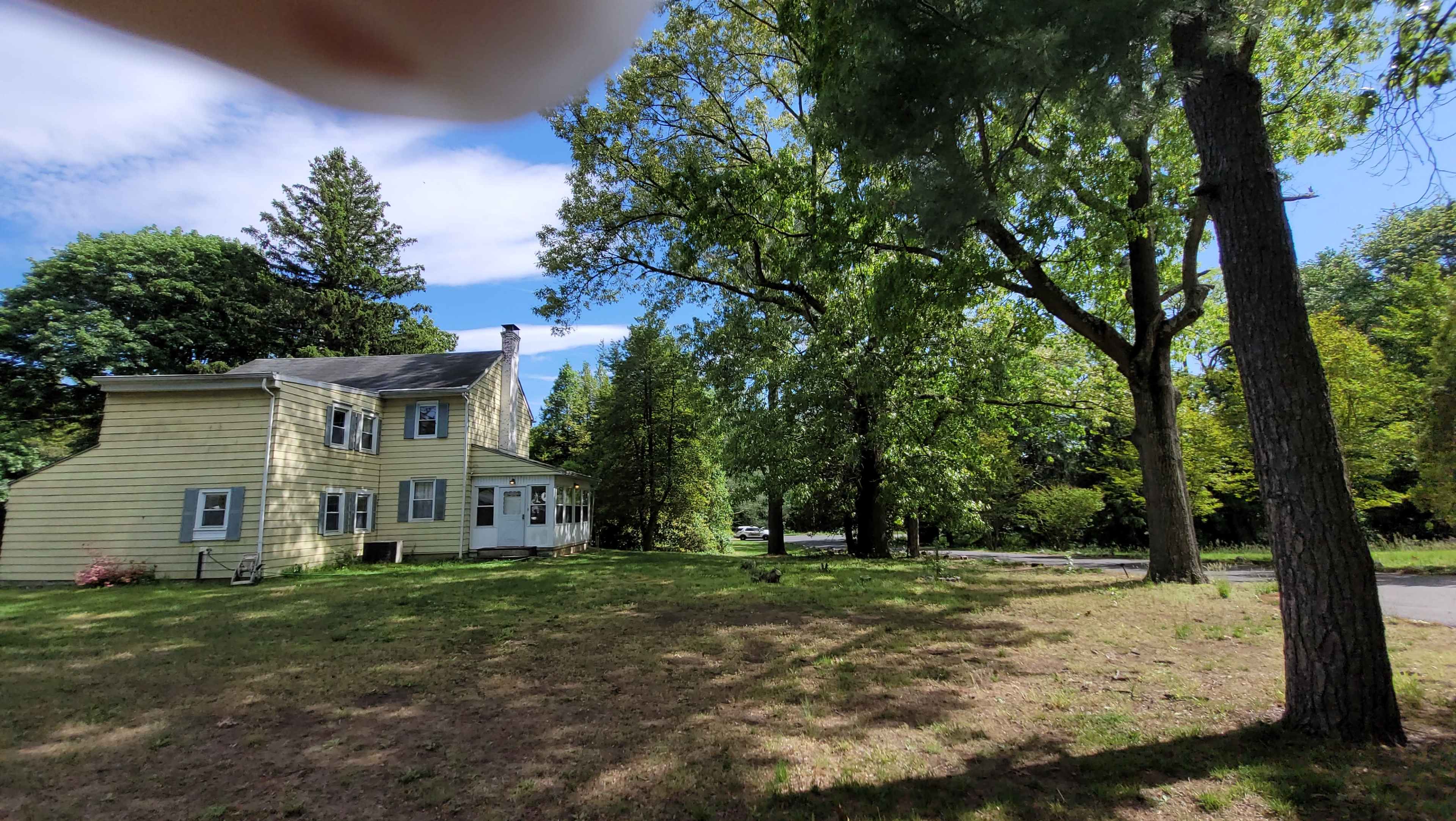 A yellow two-story house is situated on a grassy area surrounded by tall trees under a partly cloudy sky.