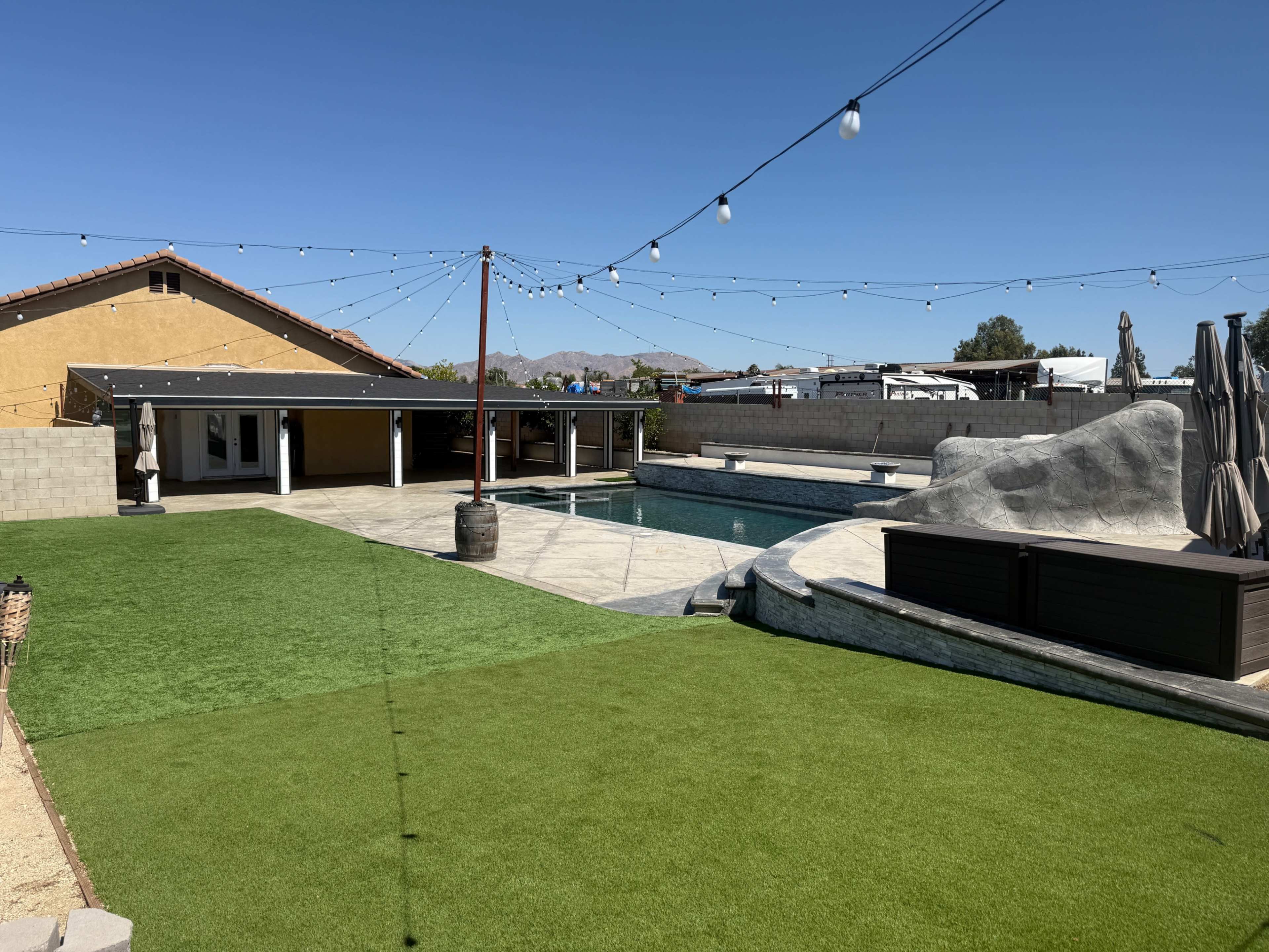 The image shows a residential backyard featuring a swimming pool, a patio area with seating, a rock formation, and synthetic grass under a clear blue sky.