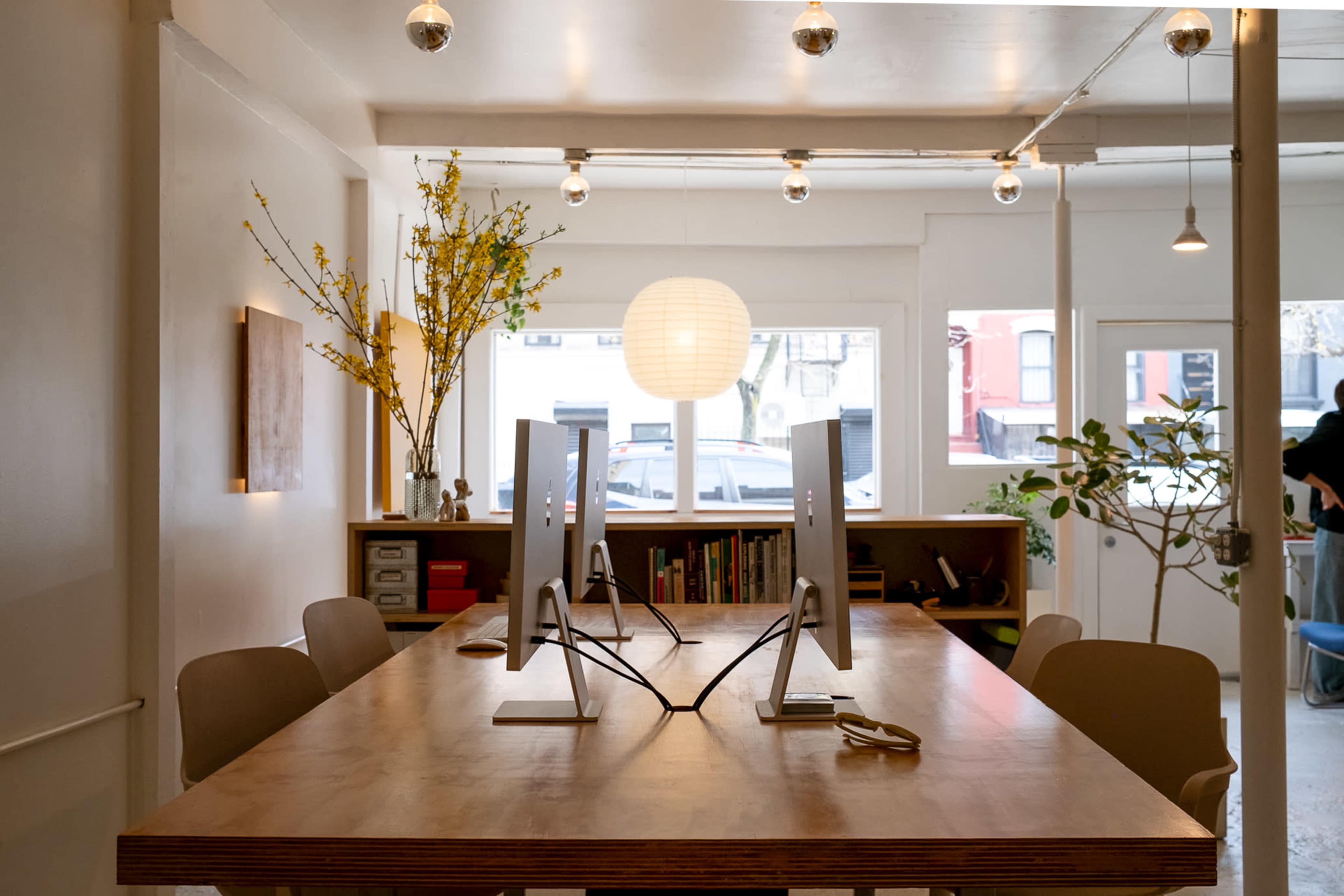 The image shows a modern office space with a large wooden table, two computer monitors on stands, and soft lighting provided by pendant bulbs and a paper lantern.