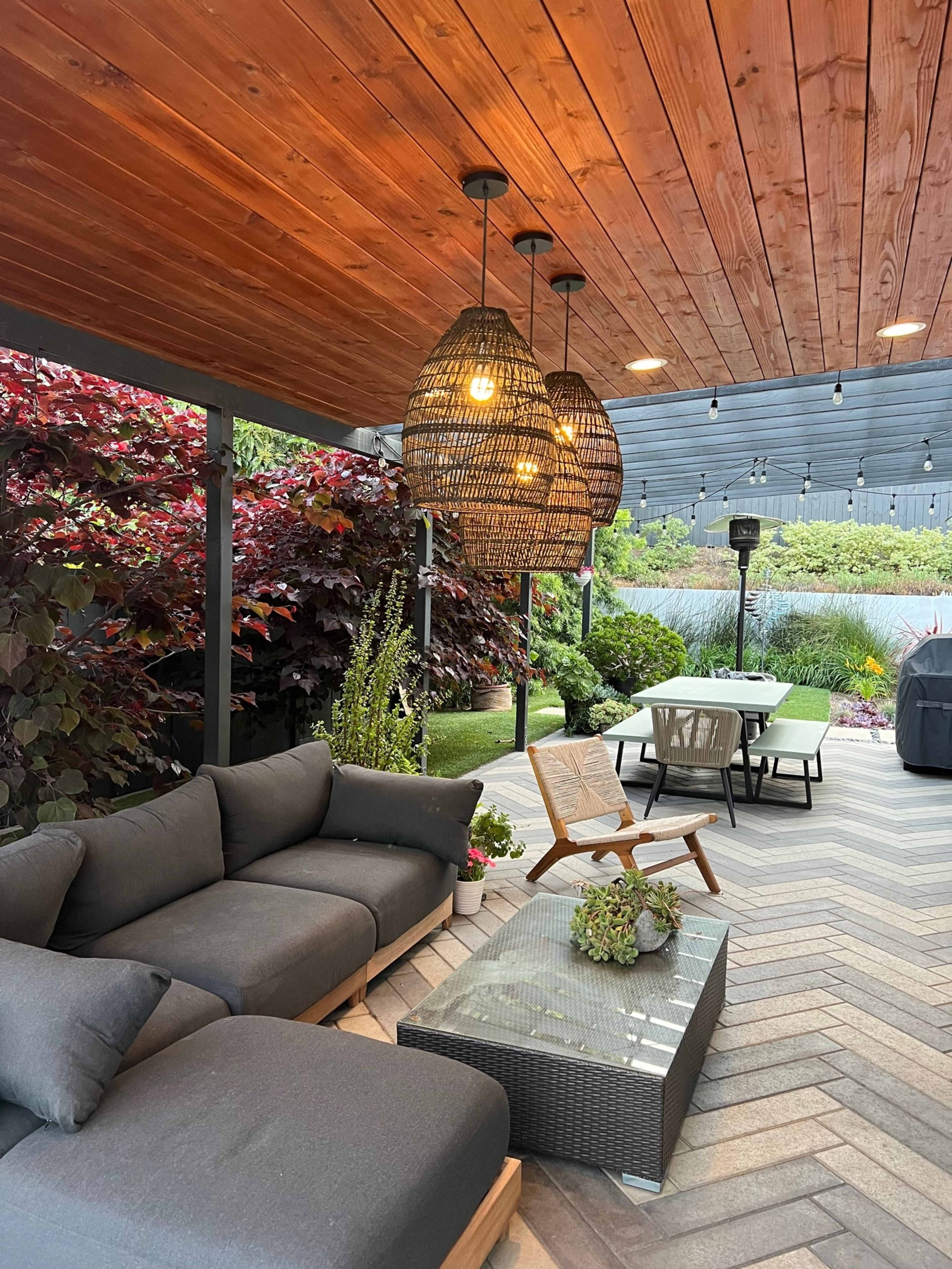 A cozy outdoor patio area featuring a sectional sofa, a glass coffee table, and three pendant lights hanging from a wooden ceiling, surrounded by lush greenery and a dining table in the background.