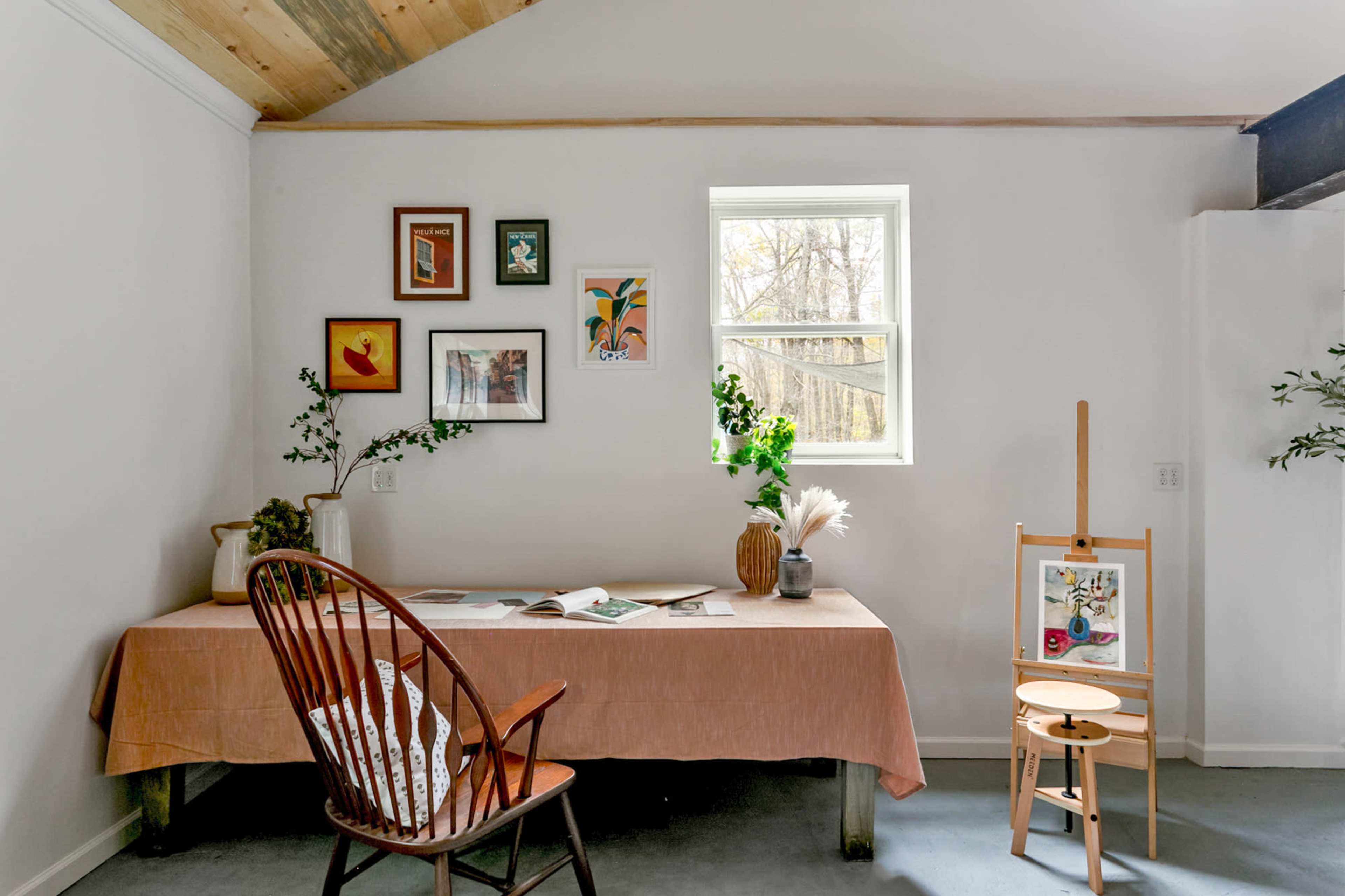 A room features a table covered with a cloth, surrounded by a chair, an easel with artwork, and framed pictures on the wall beside a window.