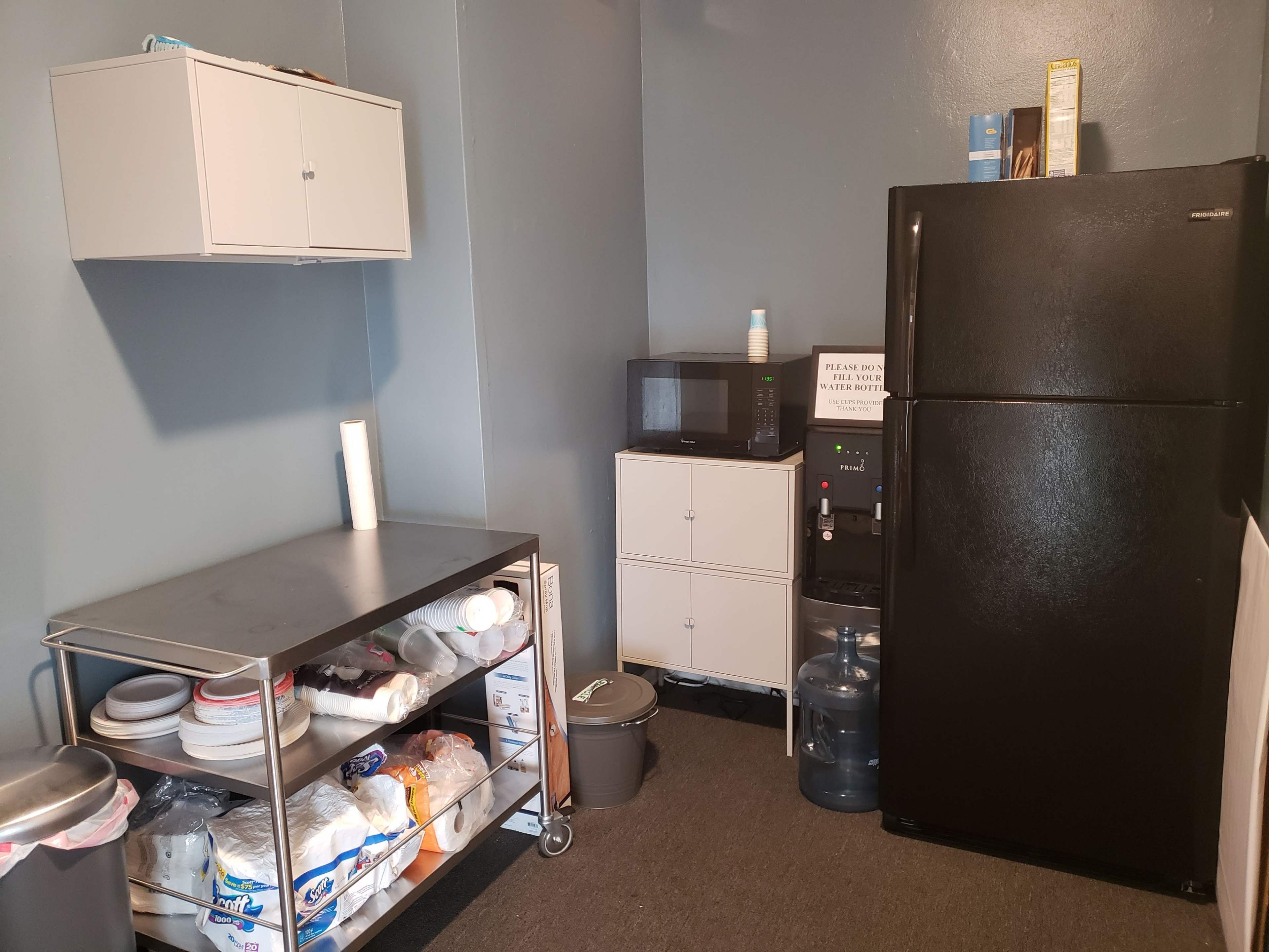 A small kitchen area featuring a black refrigerator, a microwave on a shelf, a metallic table with plates and containers, and a water dispenser.