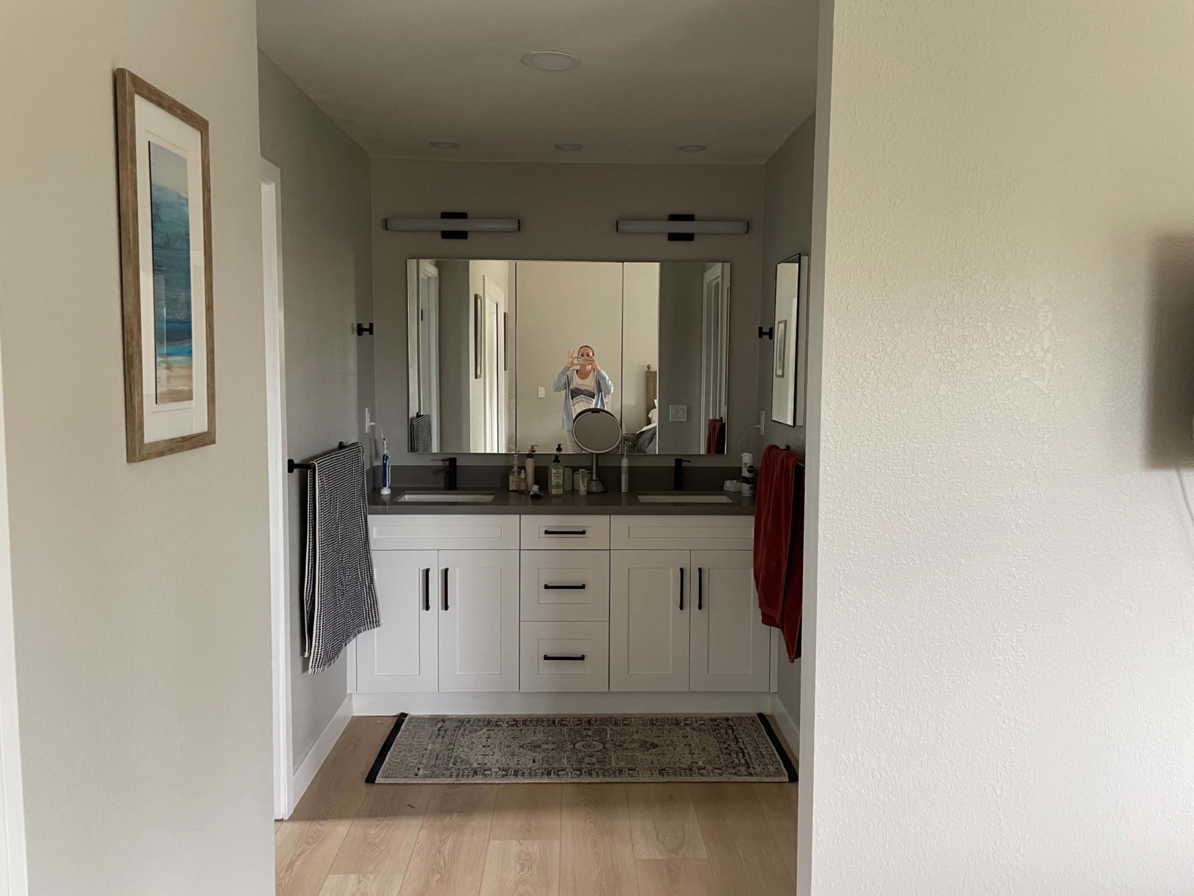 The image shows a modern bathroom with two sinks, a large mirror, and light-colored walls, featuring a rug and towels hanging on the walls.
