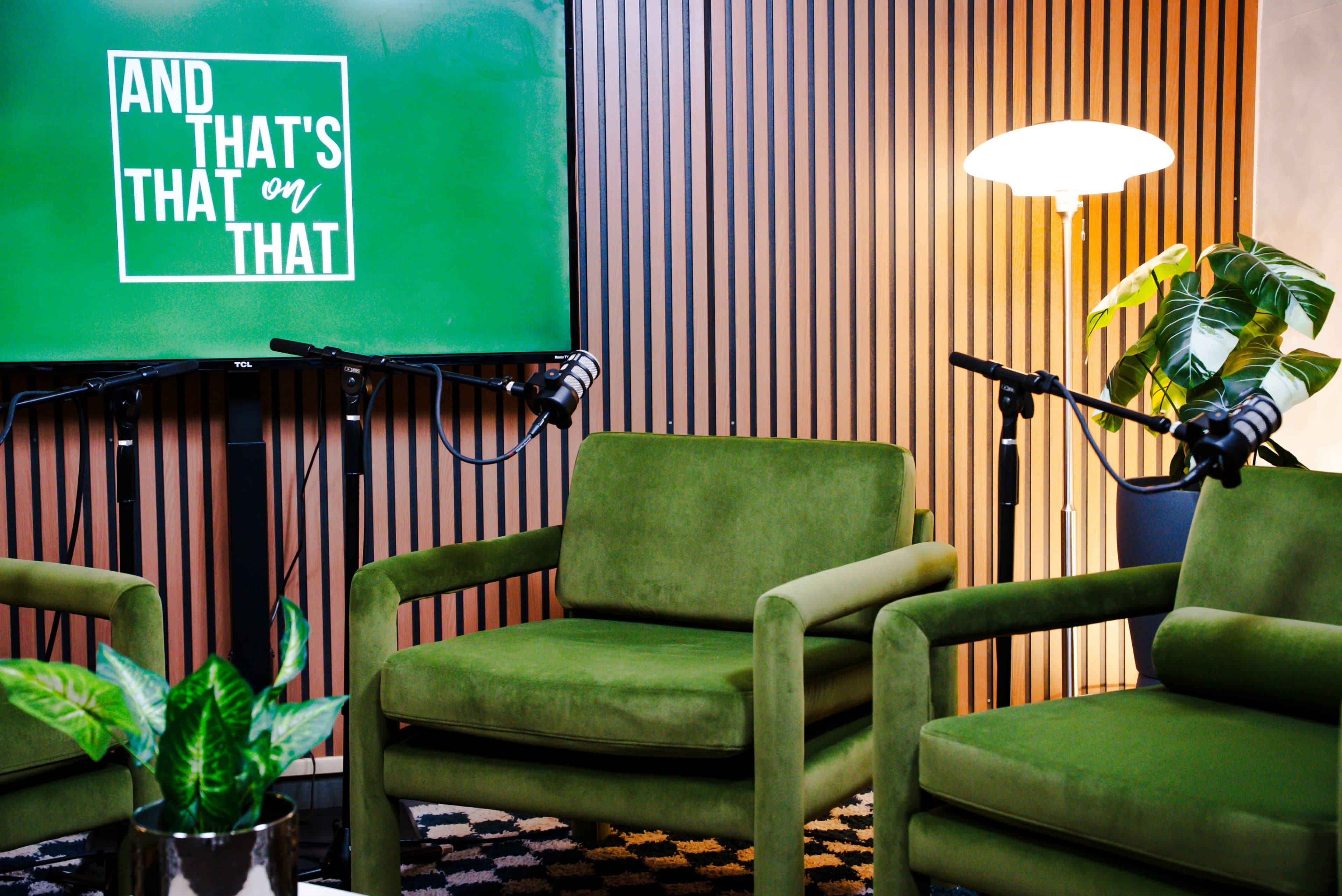 The image shows a modern interior setup with two green armchairs, a plant, and microphones positioned in front of a screen displaying the text "AND THAT'S on THAT."