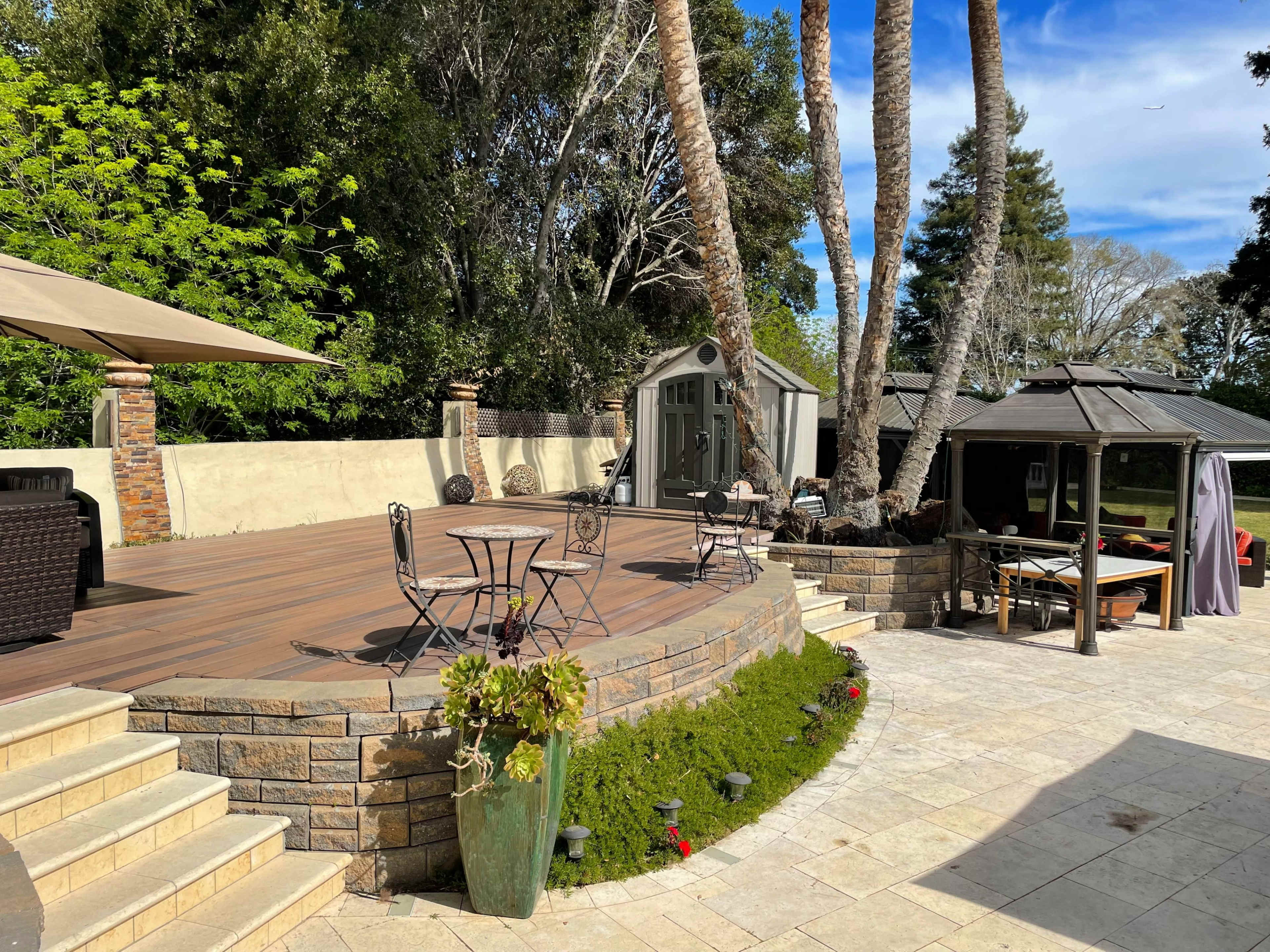 A landscaped outdoor patio with a wooden deck, several seating arrangements, palm trees, and a gazebo.