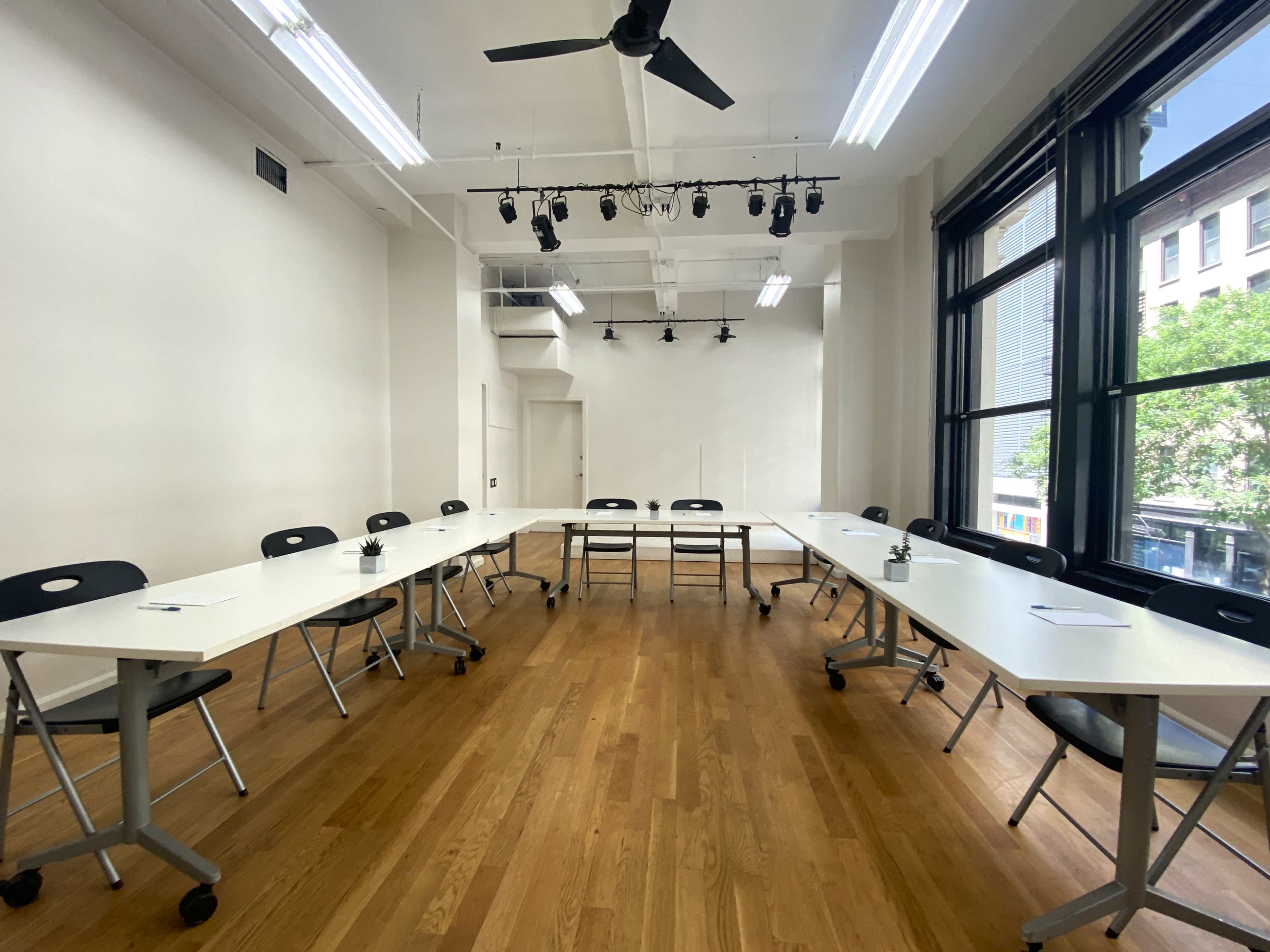 A modern conference room features a U-shaped arrangement of white tables and black chairs, with large windows allowing natural light to enter.