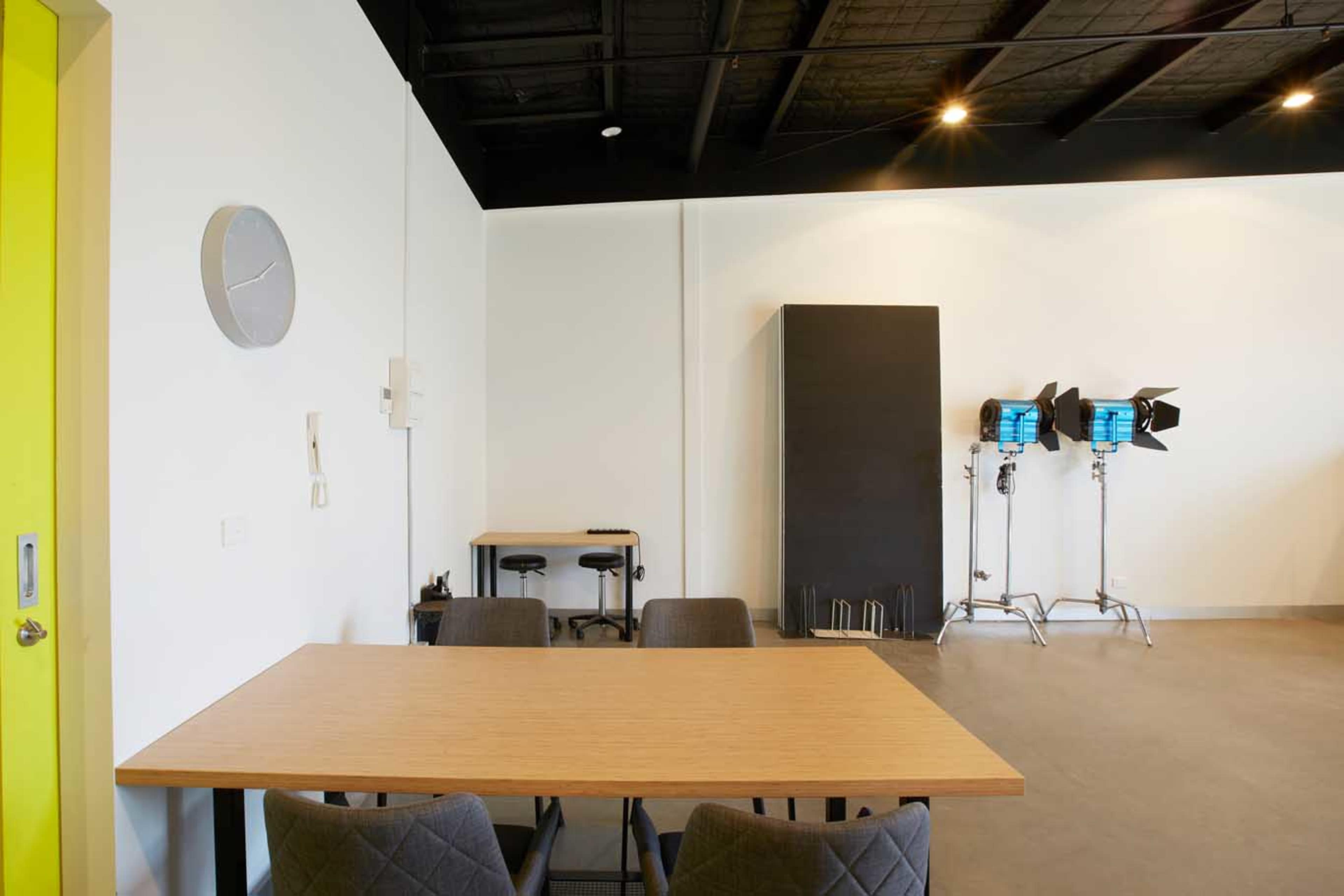 A bright, minimalist room with a wooden table and chairs, a clock on the wall, and professional lighting equipment in the corner.