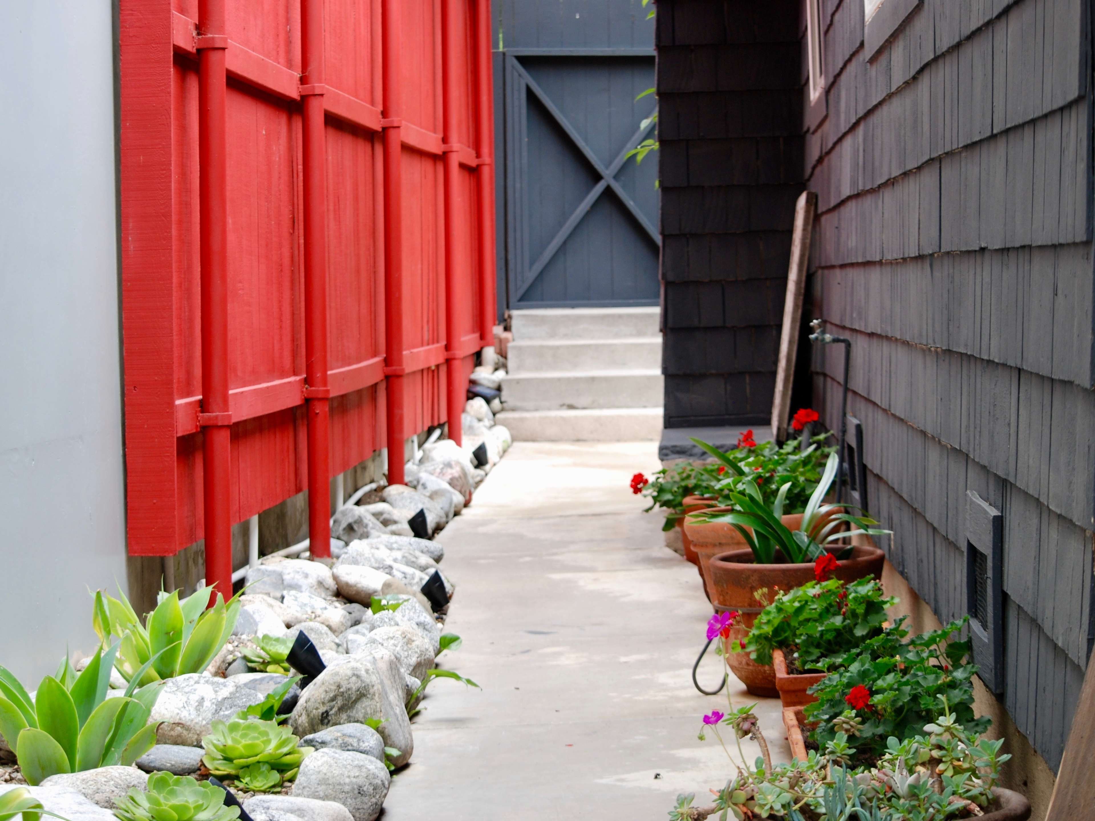 A narrow pathway is flanked by a red wooden fence on one side and a dark shingled wall on the other, with various potted plants and rocks along the edges leading to a gray door at the end.