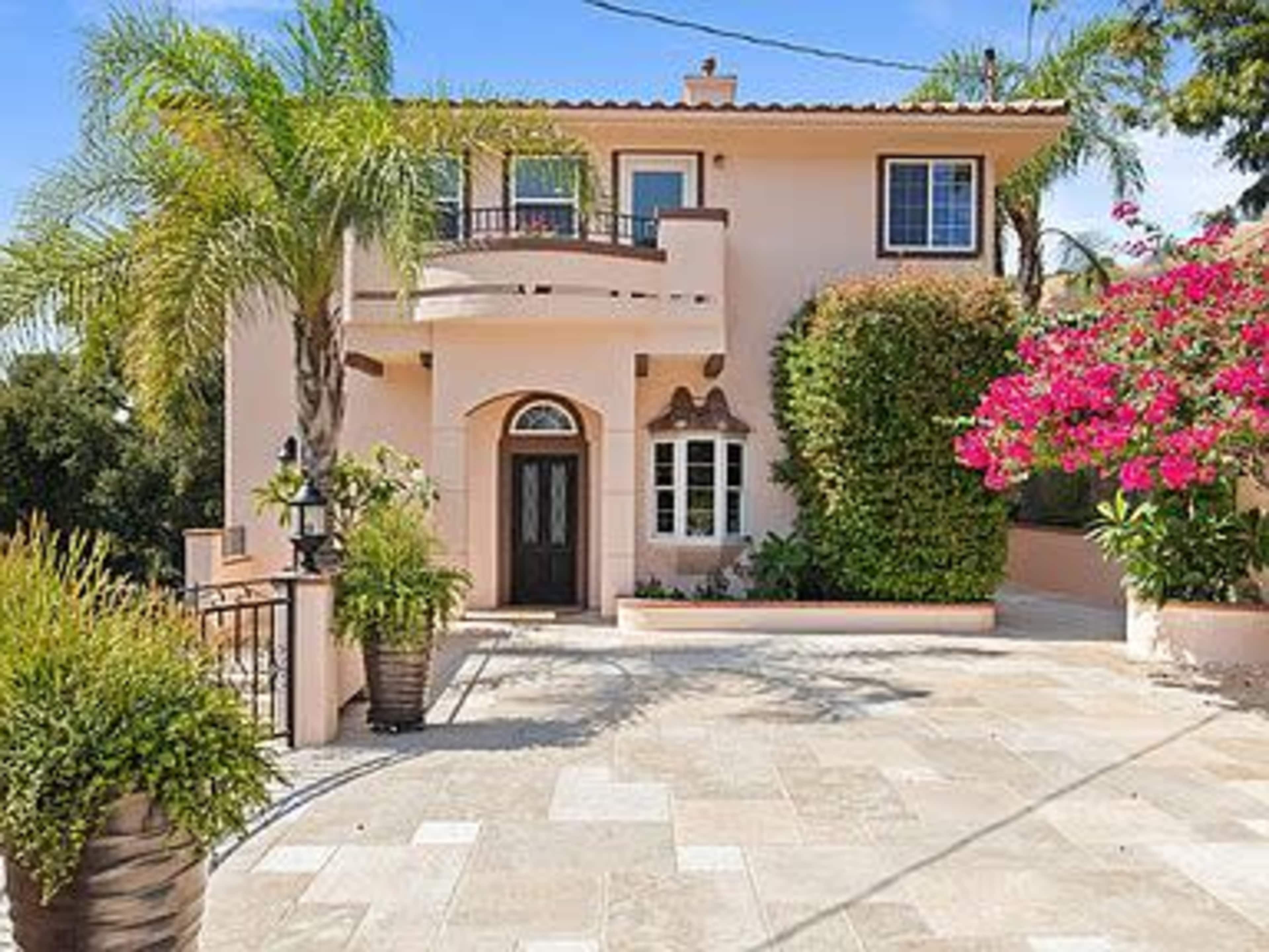 A two-story house with a pink exterior, surrounded by palm trees and vibrant bougainvillea, features a front entrance and a stone driveway.
