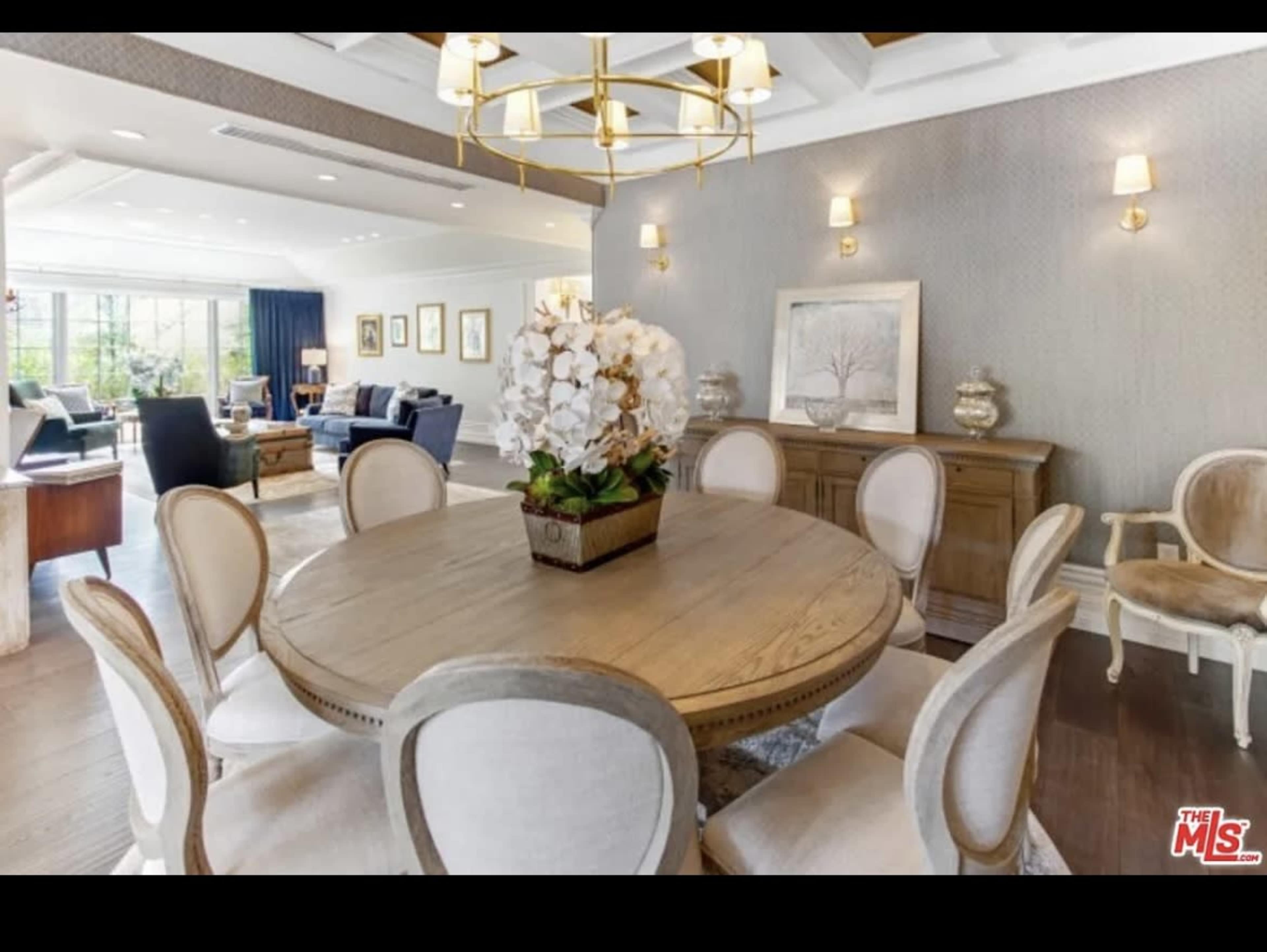 A modern dining room featuring a round wooden table with eight chairs, adorned with a centerpiece of white orchids, and large windows that provide natural light.
