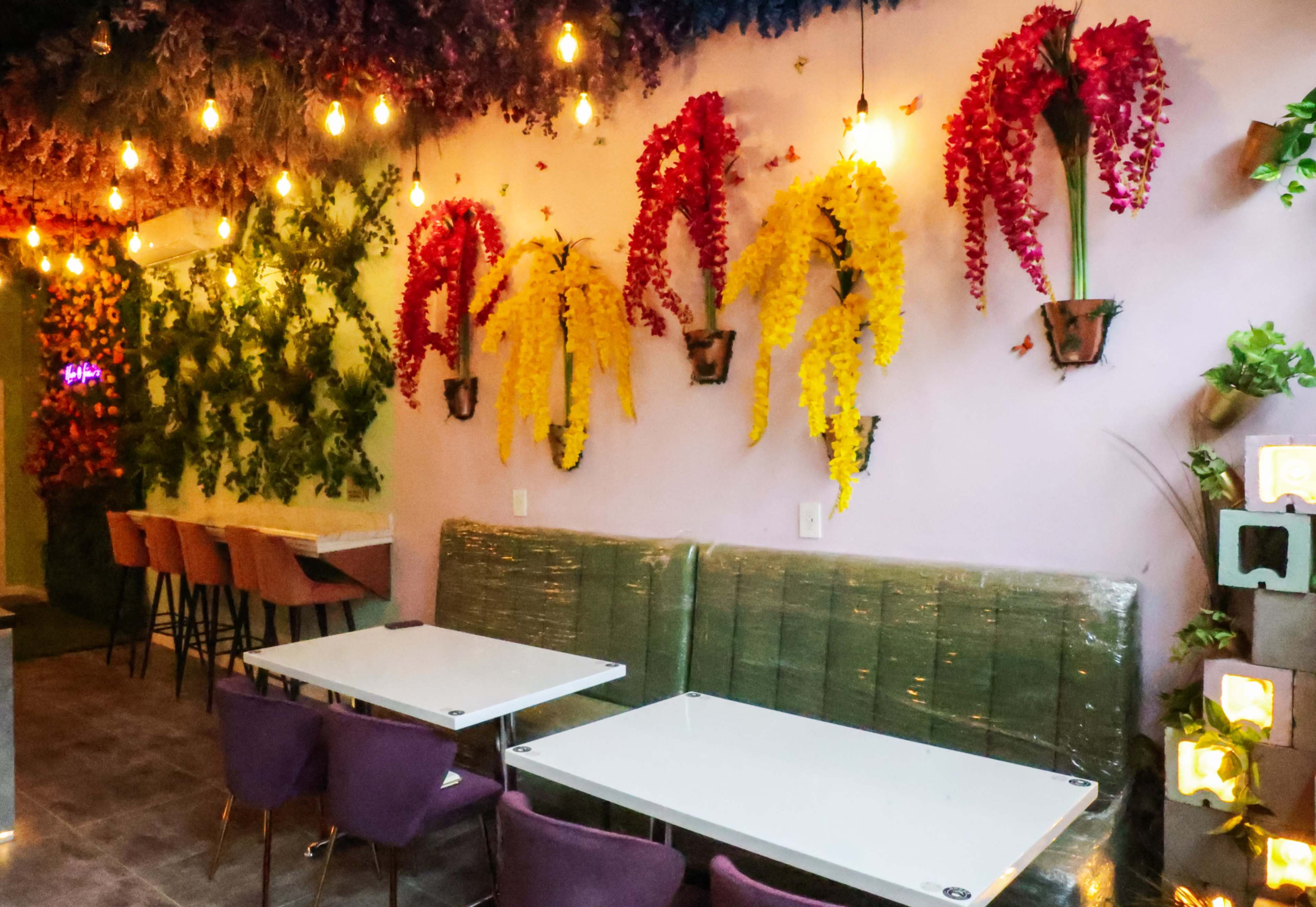 A café interior adorned with colorful artificial flowers on the walls, complemented by a row of seating and tables.