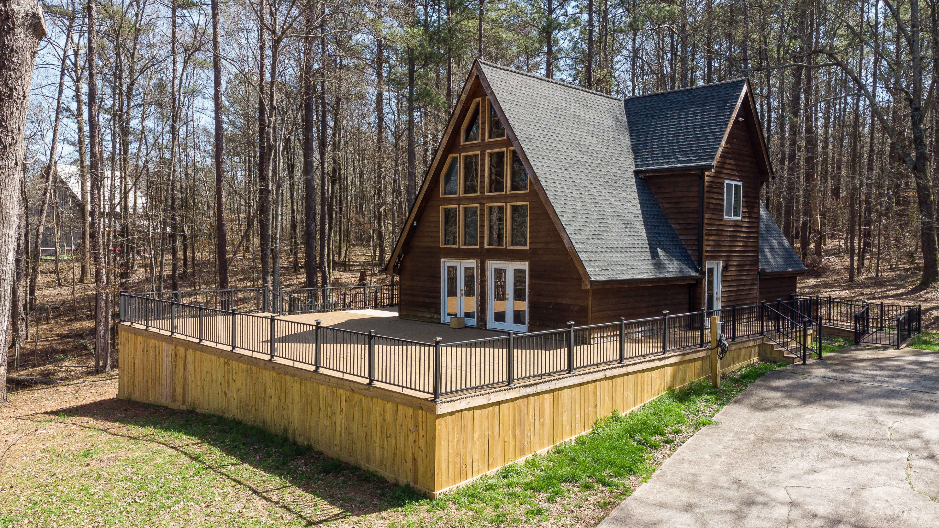 A brown A-frame house with large windows is surrounded by trees, featuring a large deck and a paved driveway.
