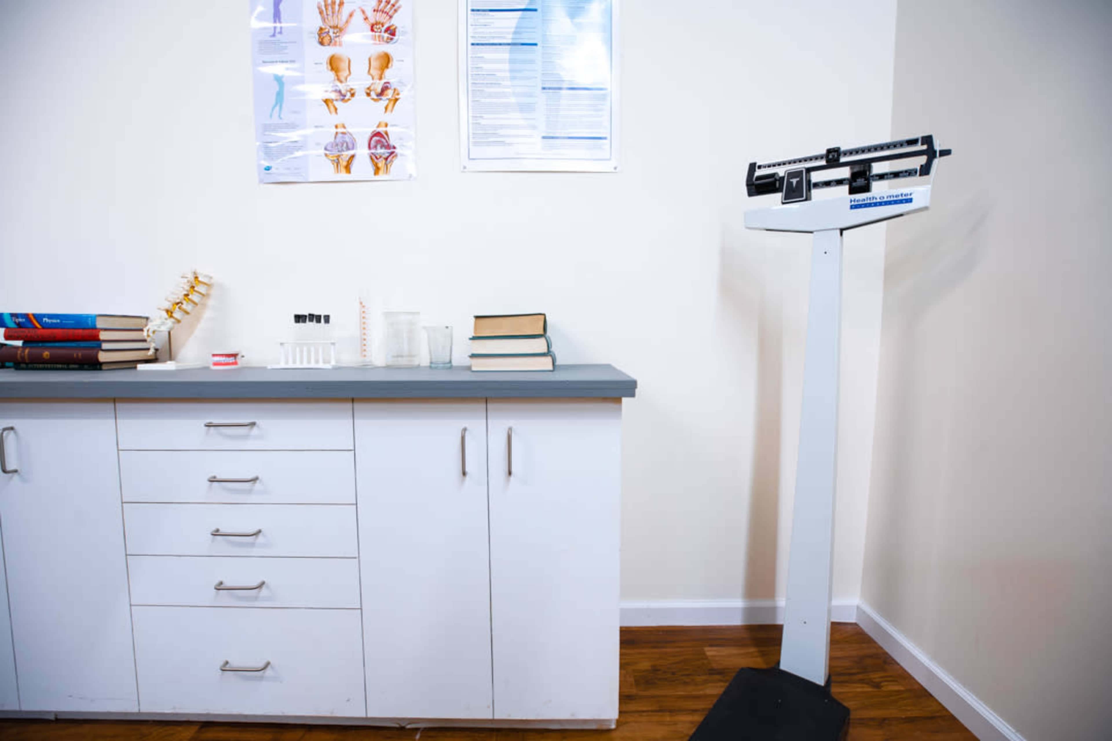 A medical examination room features a weighing scale next to a cabinet with books and medical supplies on a countertop.