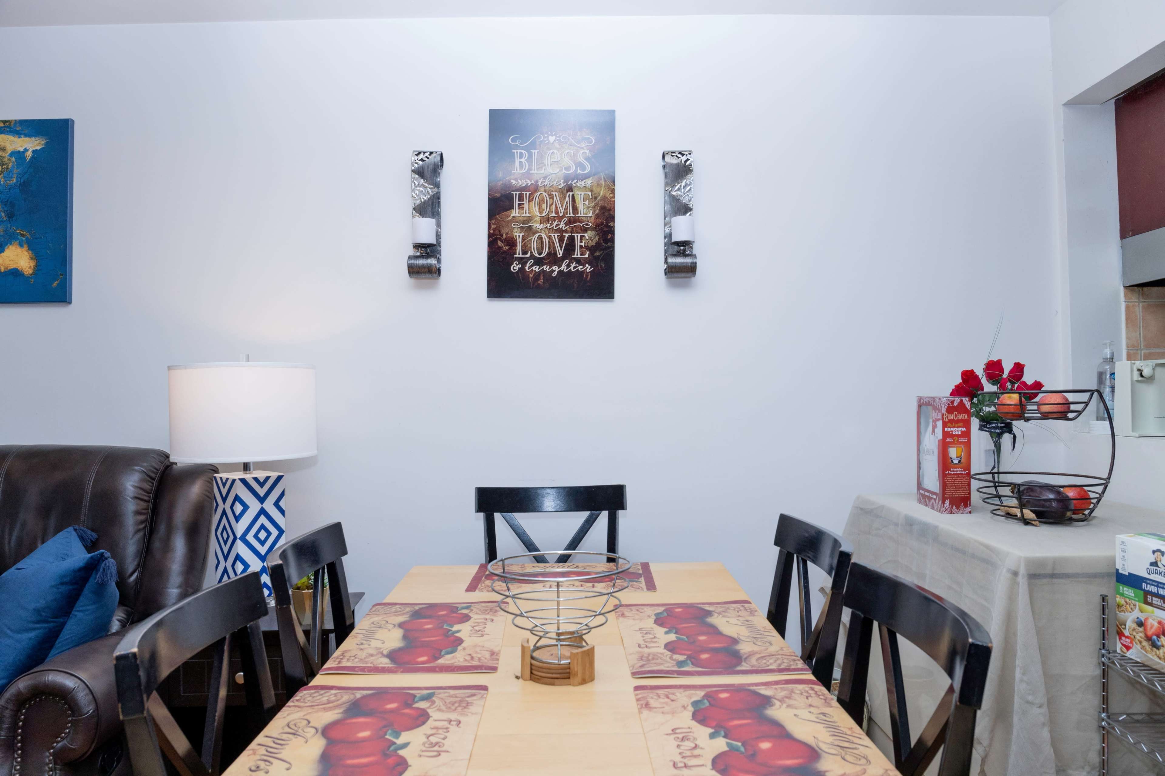 The image shows a dining area with a wooden table set with a fruit-themed tablecloth, surrounded by black chairs, and wall decorations above.