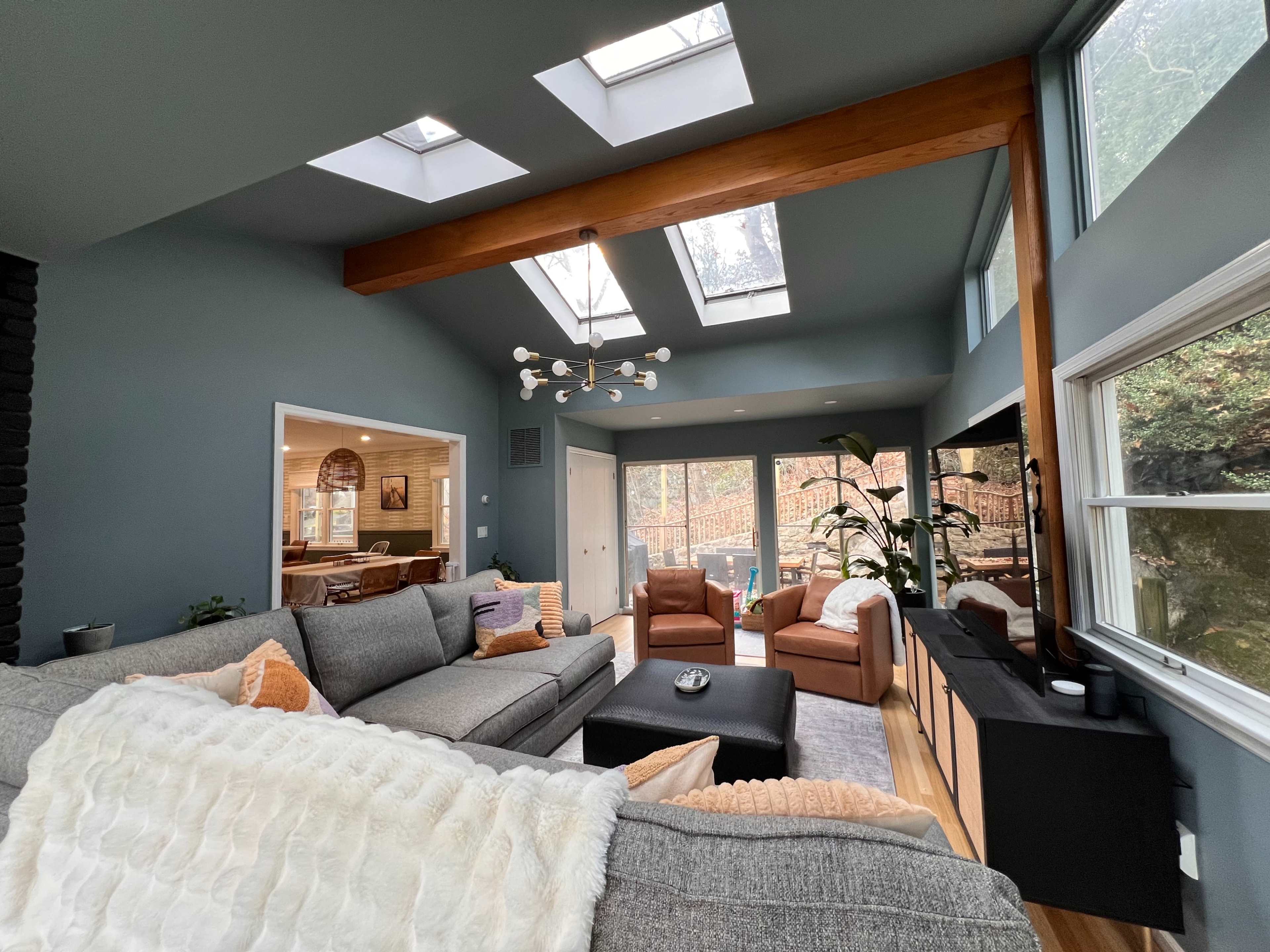 A modern living room with large windows, skylights, and a mix of comfortable furniture including a gray sectional sofa, two brown armchairs, and a coffee table.