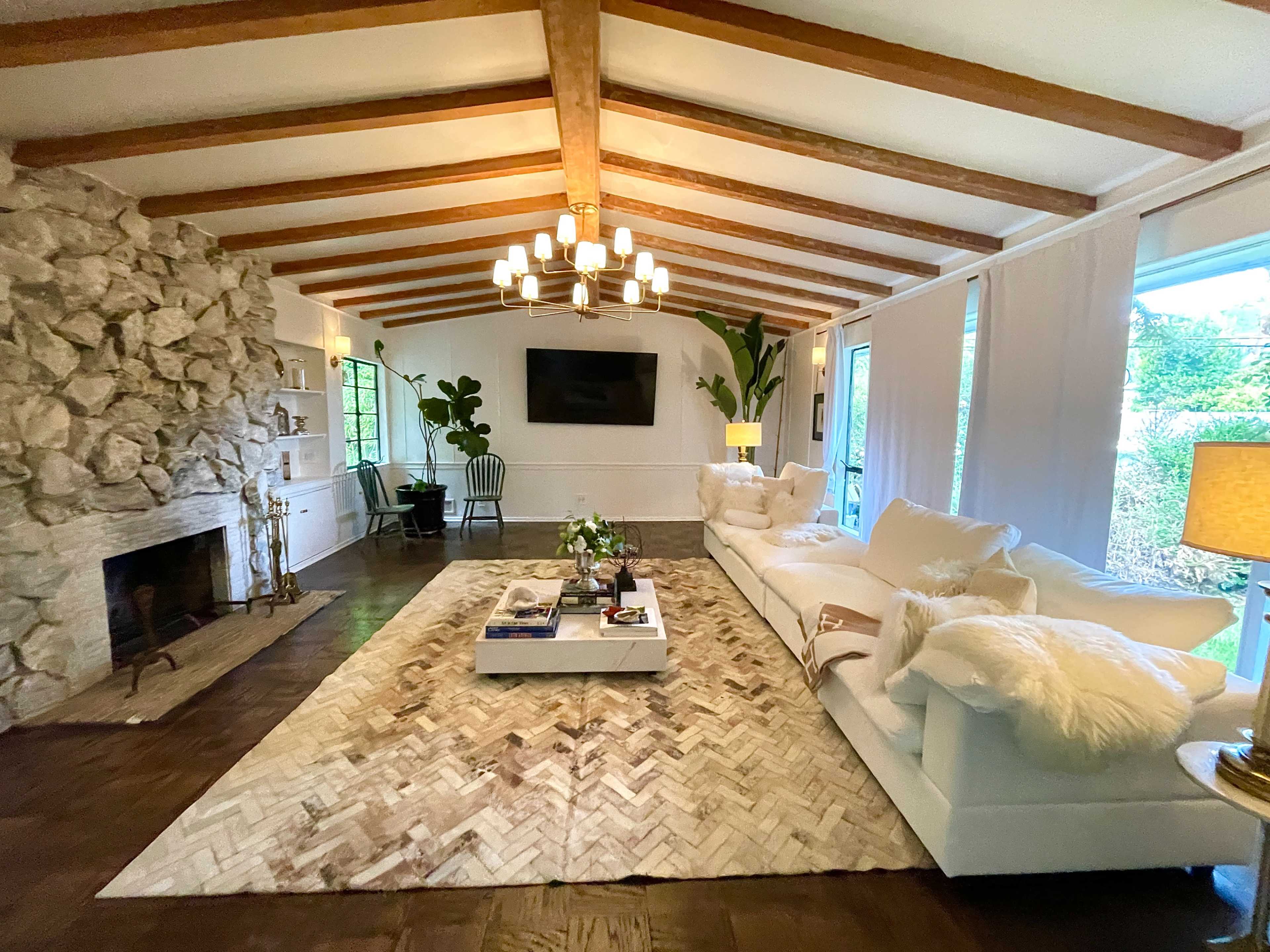 The image shows a spacious living room with a stone fireplace, a large white sofa, a coffee table on a patterned rug, and plants near the windows.