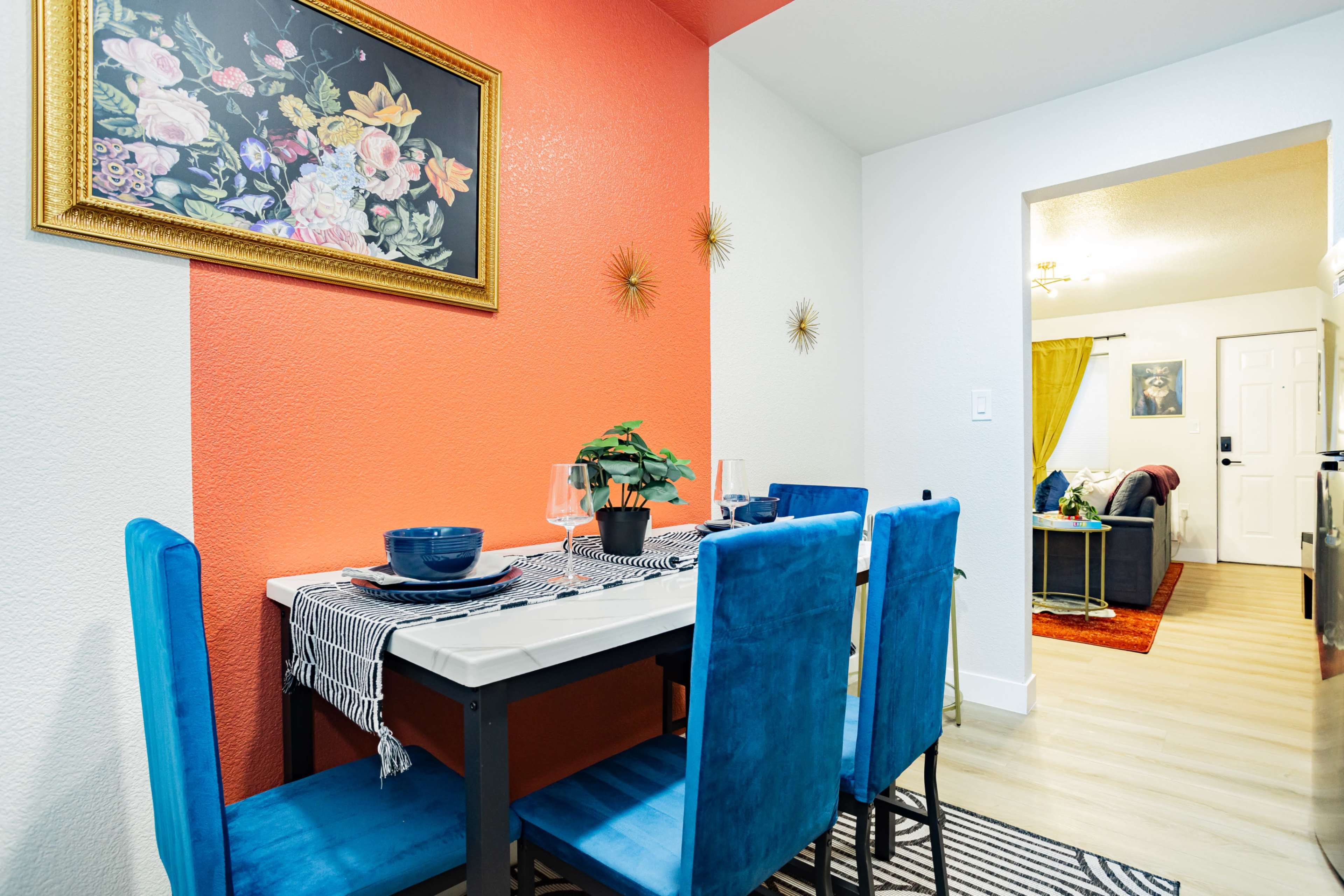 A dining area features a rectangular table set for two with blue velvet chairs, surrounded by an orange accent wall and a doorway leading to a living space.