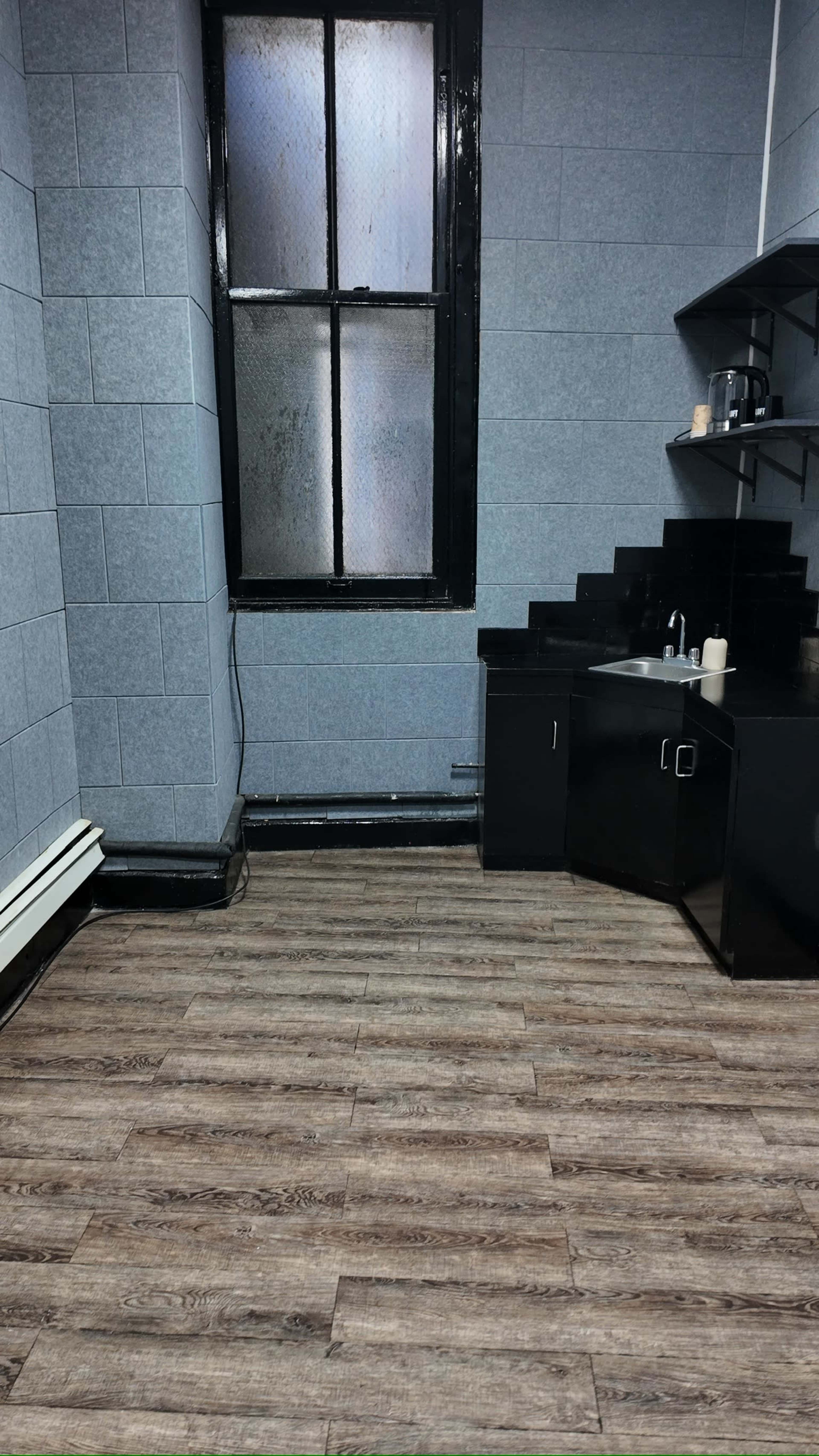 The image shows a sparsely furnished kitchen area featuring gray walls, a window with condensation, and a black countertop with a sink and a few kitchen appliances.