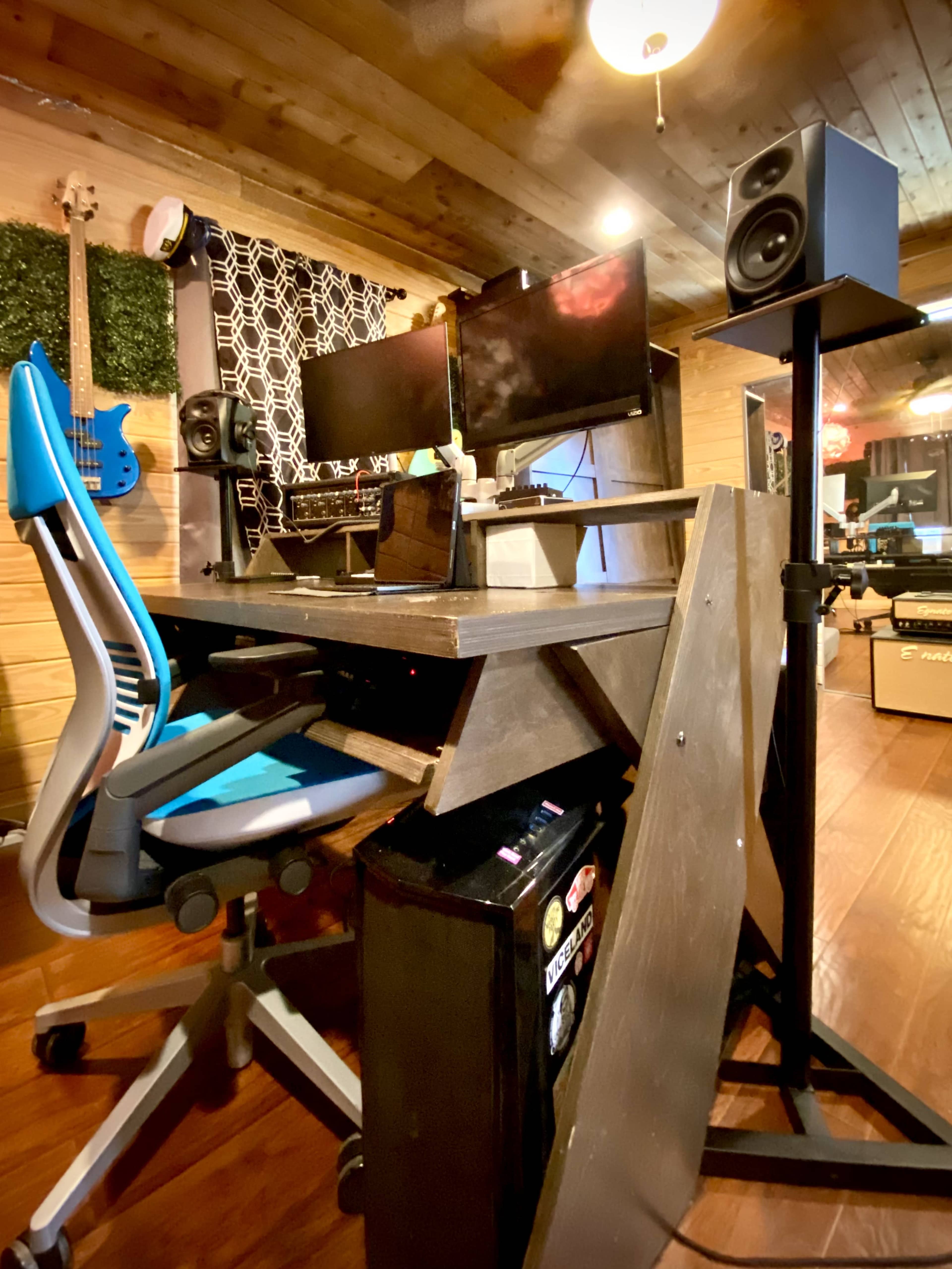 A desk setup featuring dual monitors, a chair, and a speaker stand in a wooden room.