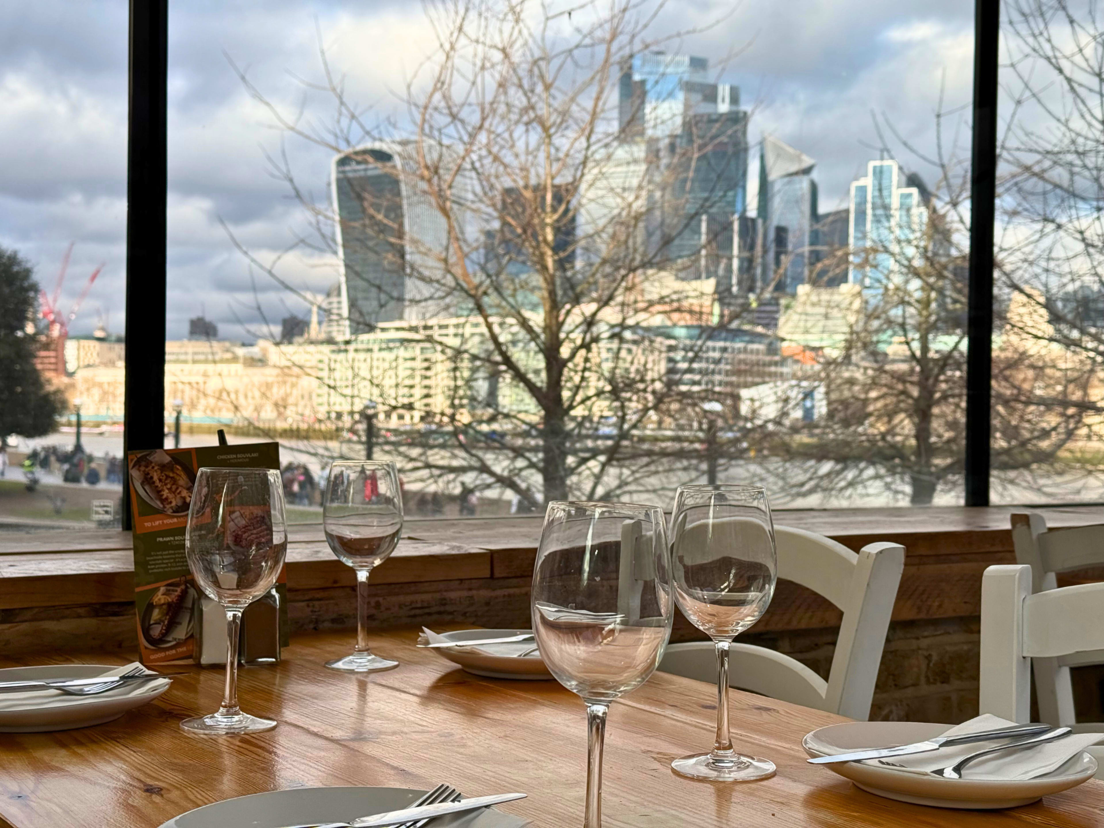 The image shows a dining table set with glassware and plates, overlooking a cityscape featuring modern buildings across a river.