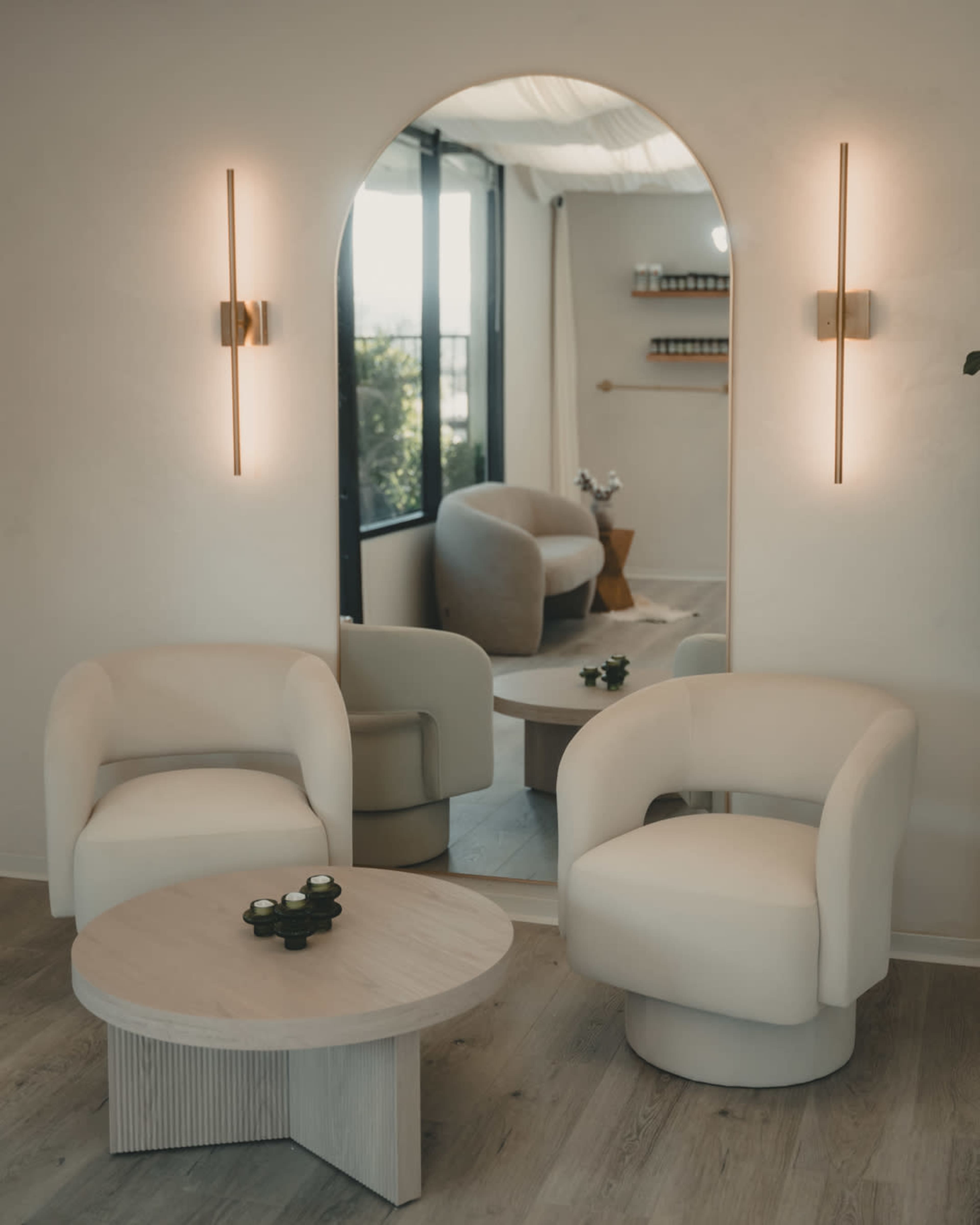 The image shows a modern, minimalist interior featuring two light-colored armchairs, a round wooden table, and a large mirror reflecting part of the room.
