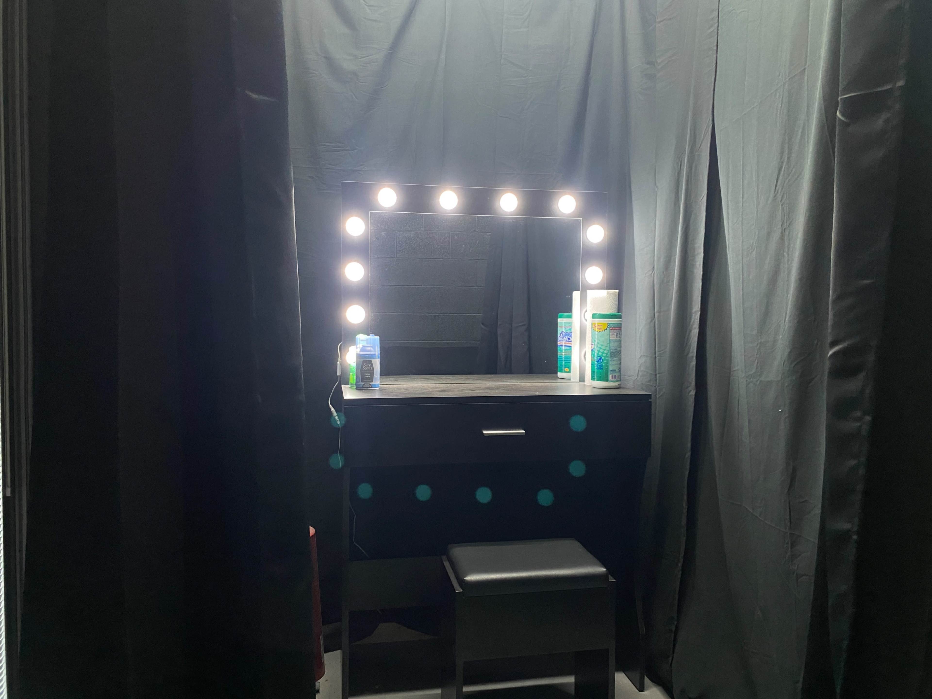 A well-lit vanity with a large mirror and bottles on it is backed by black curtains.