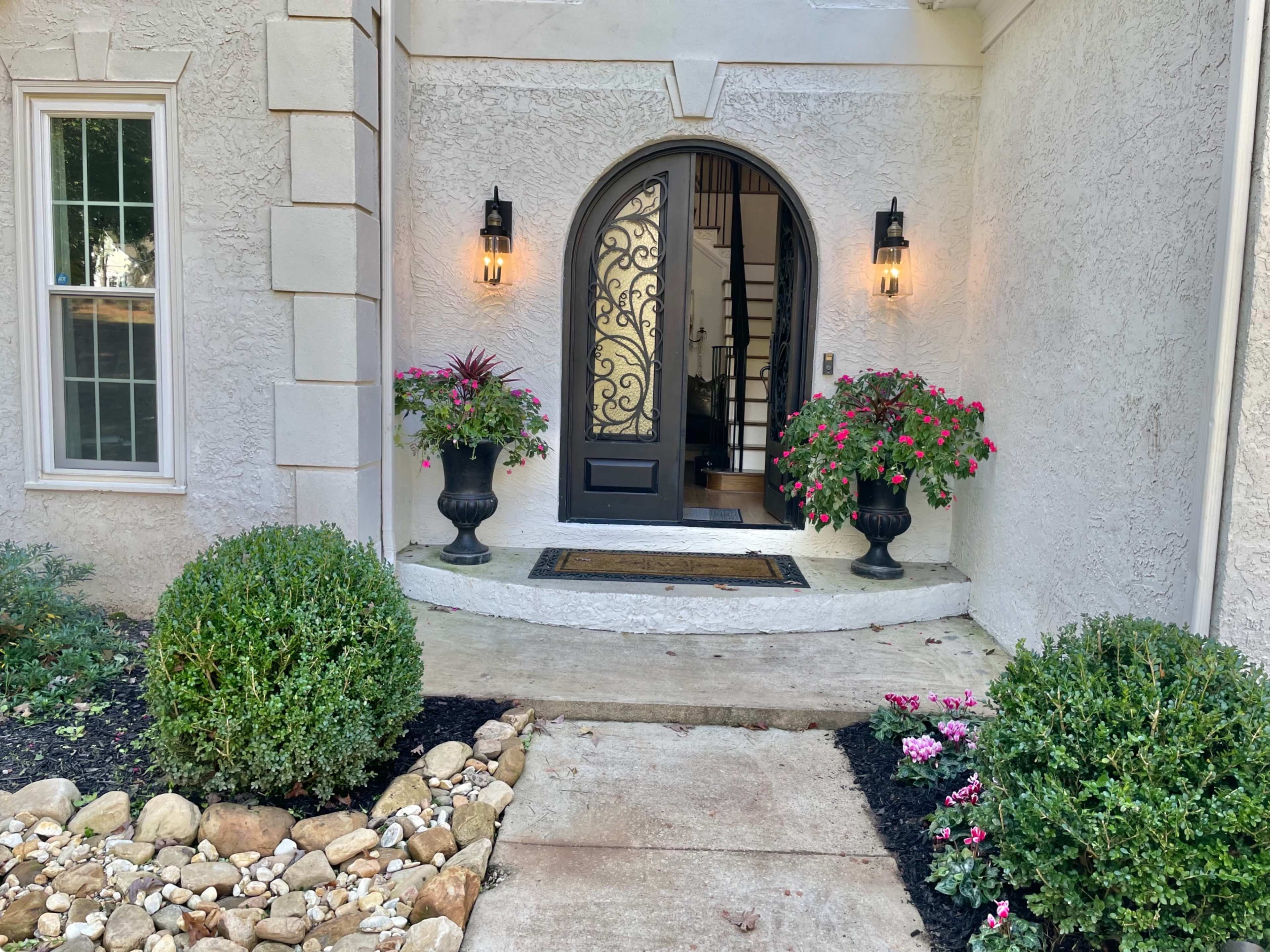A front entrance with a curved step leading to a black door framed by decorative plants in large urns, flanked by stone walls and a neat pathway.