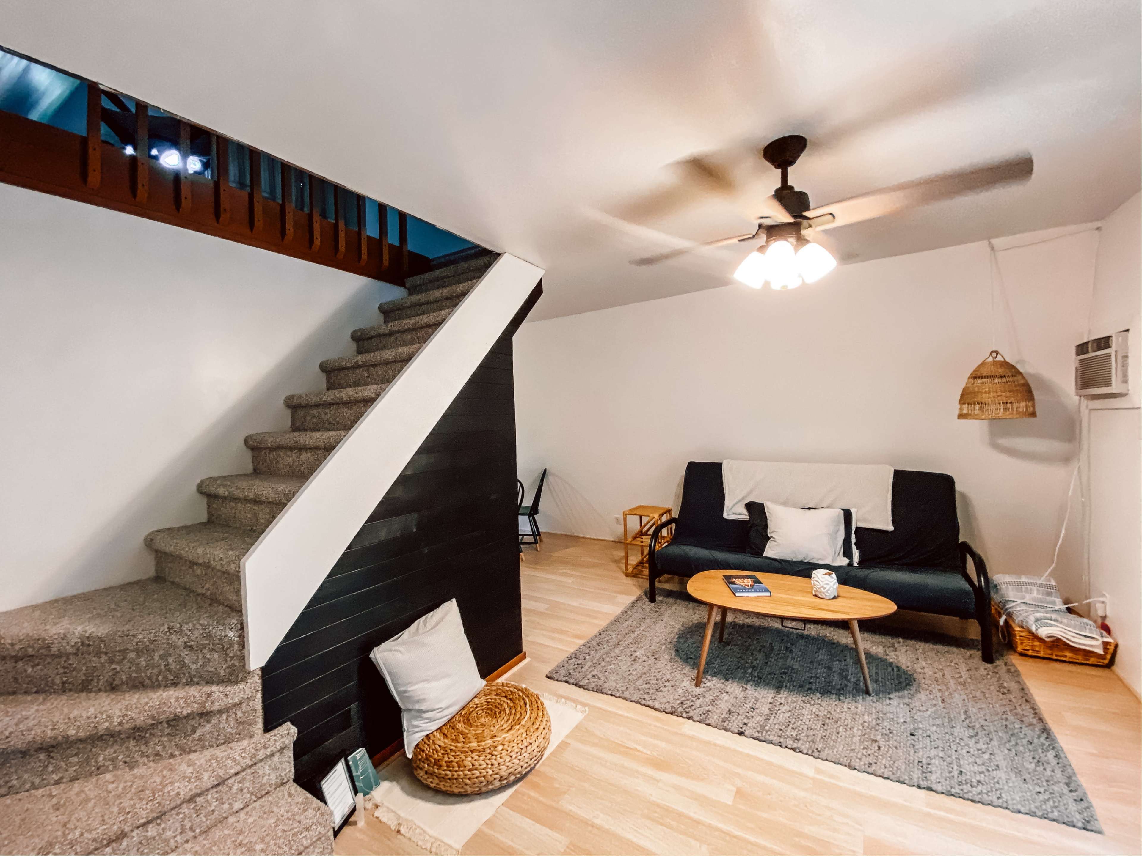 The image shows a living room with a staircase, a couch with pillows, a coffee table, and a rug on the floor.