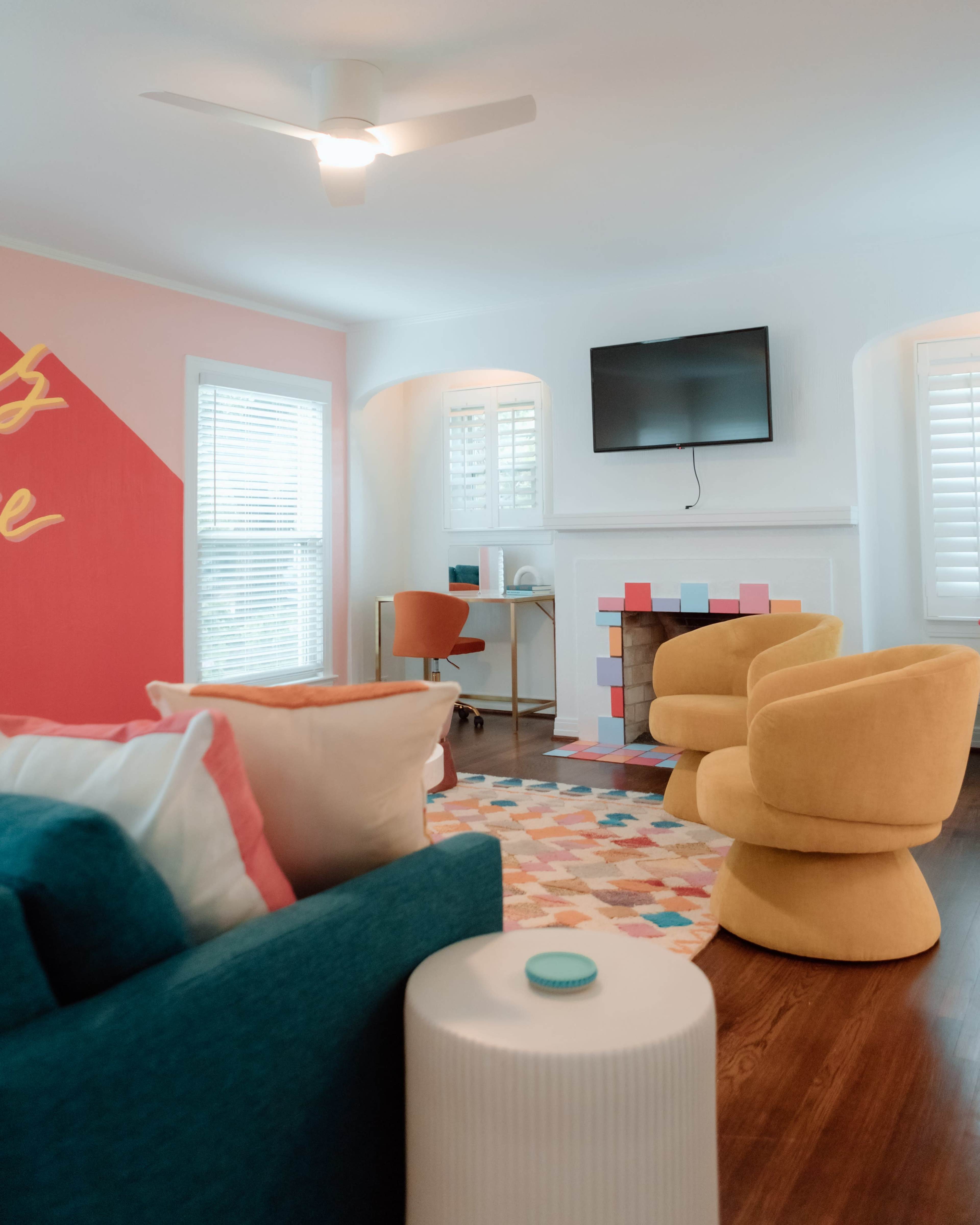 A bright living room featuring two yellow armchairs, a colorful rug, a television mounted on the wall, and a small workspace with a desk and chair.