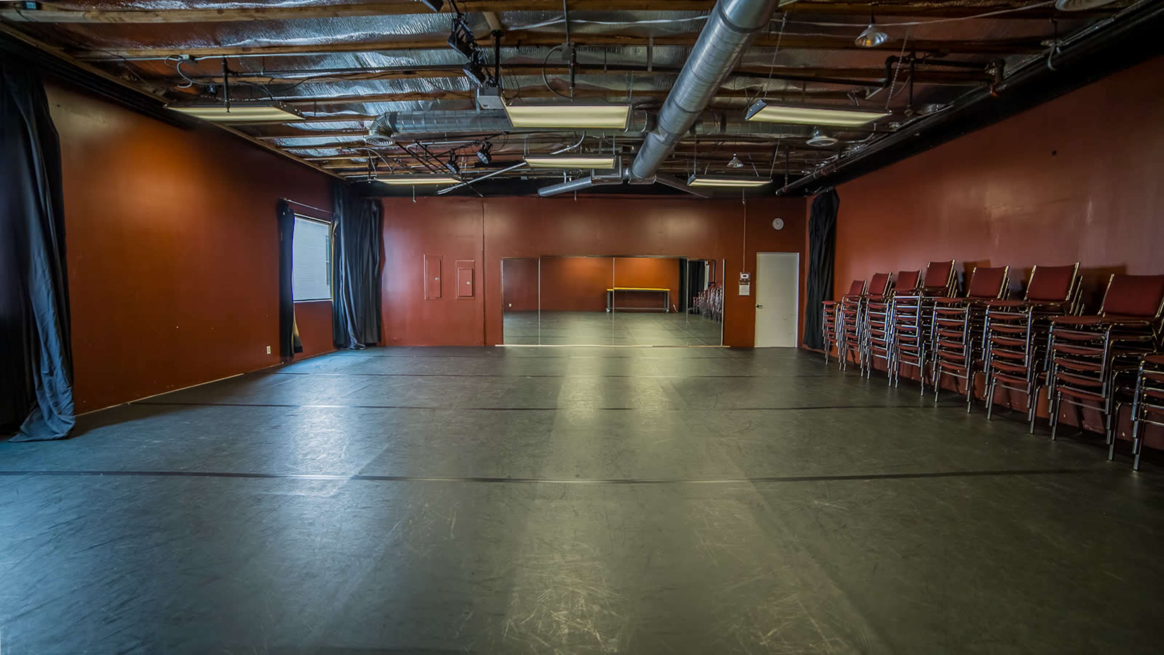 The image shows a spacious room with dark walls, a polished floor, and stacked chairs along one side.