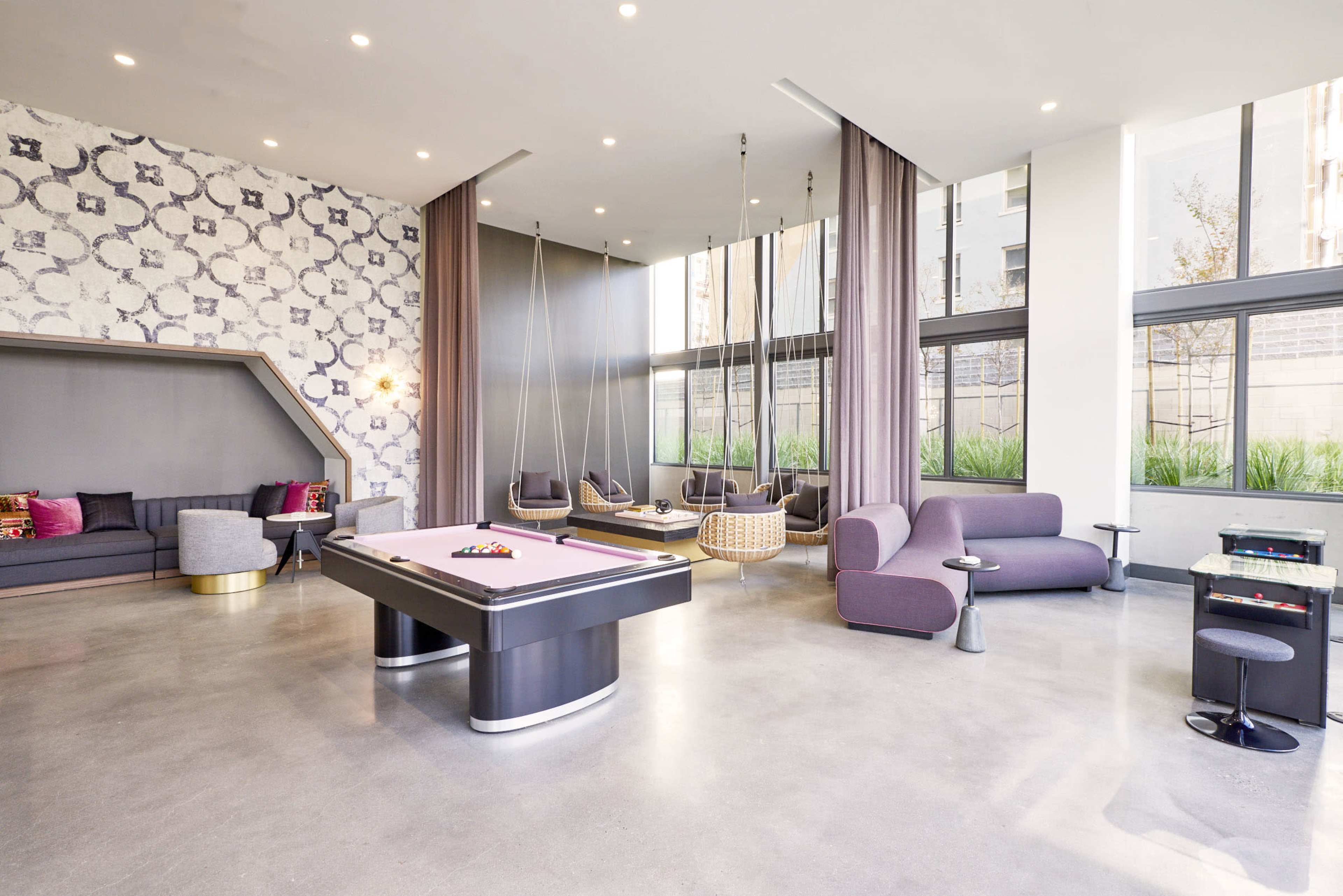 A modern lounge area featuring a pool table, hanging seating, and large windows with greenery visible outside.