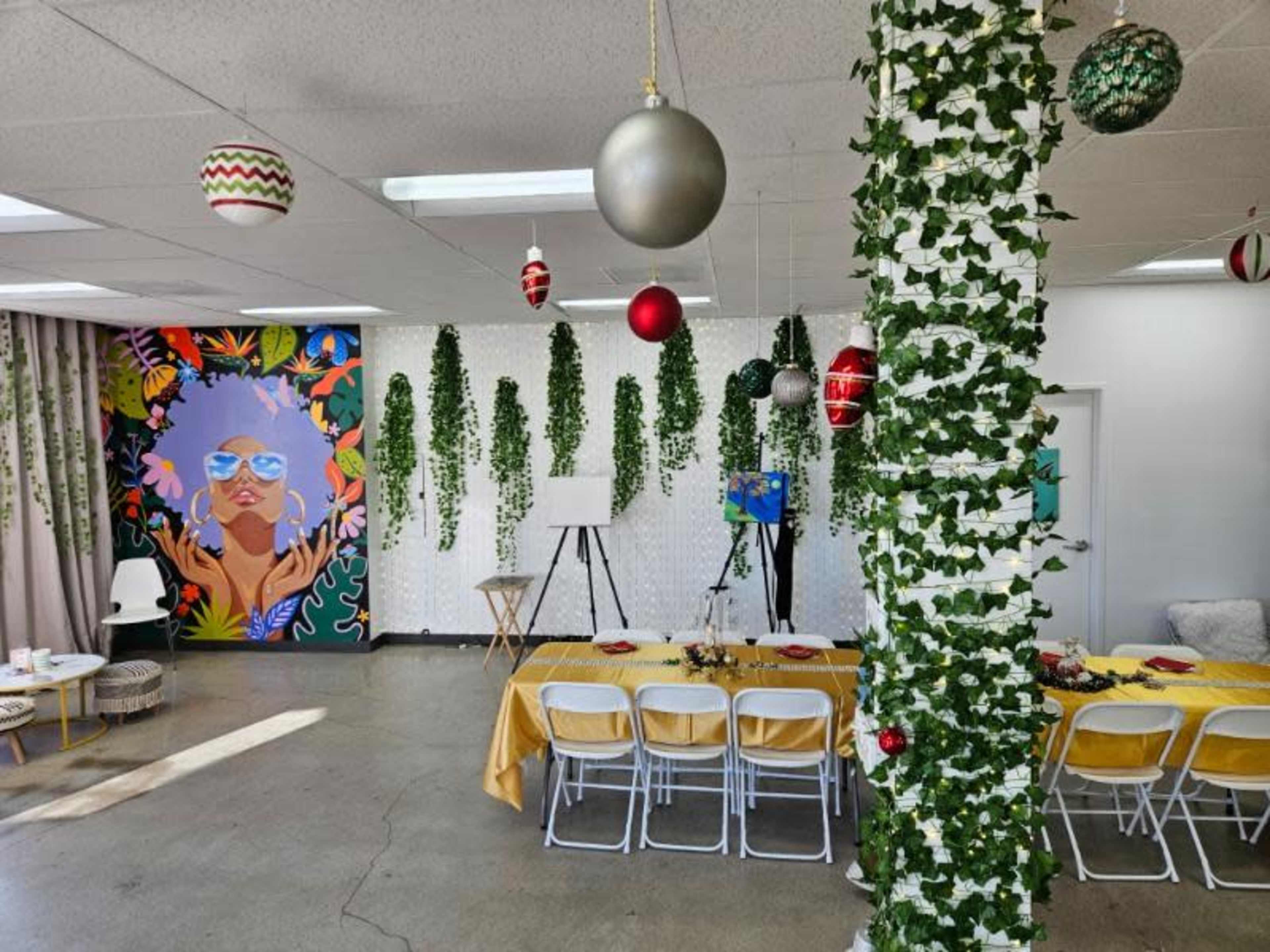 A decorated event space with a table set for a gathering, a colorful mural on the wall, and hanging ornaments among greenery.
